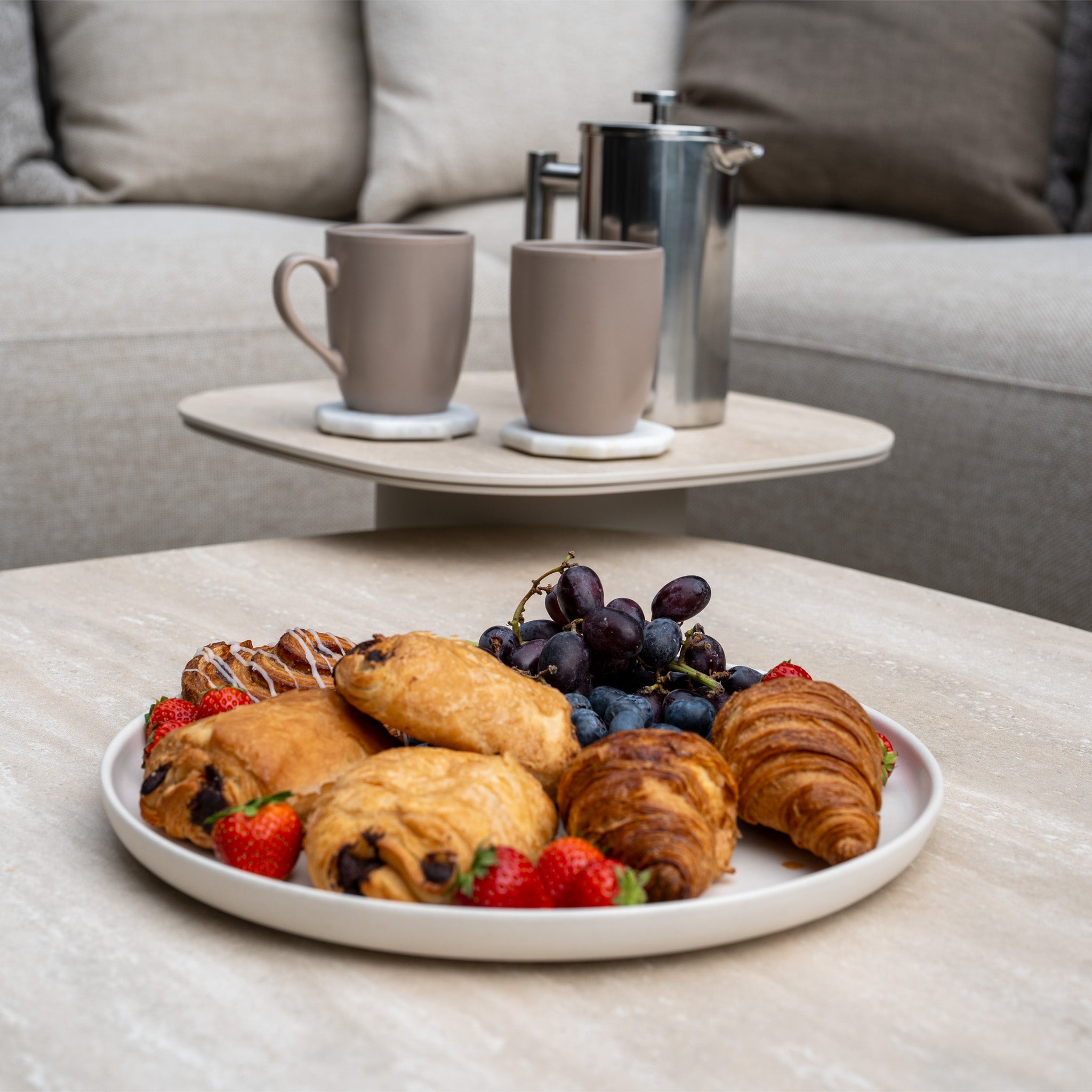A plate of pastries, strawberries, and grapes rests on a table beside the Avola Corner Sofa Set in Fawn, crafted with weather-resistant olefin—perfect for relaxed mornings with French press coffee.