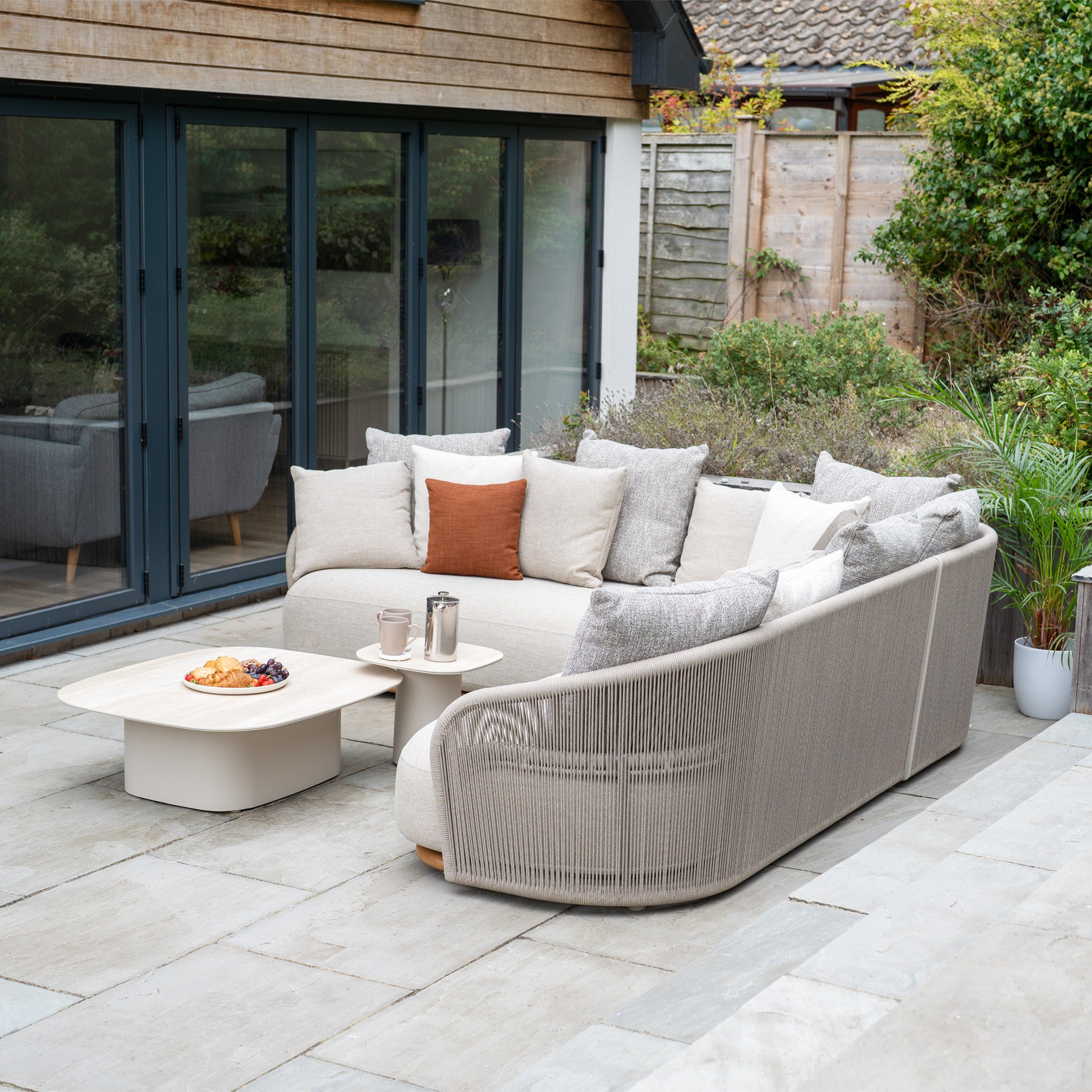 Modern outdoor patio featuring the Avola Corner Sofa Set in Fawn with weather-resistant olefin, plush cushions, a coffee table, snacks, and plants on stone flooring.