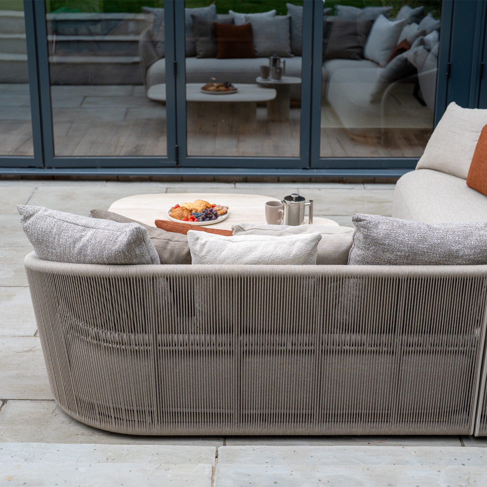 The Avola Corner Sofa Set in Fawn features weather-resistant olefin cushions and is arranged around a round table topped with pastries, cups, and a French press.