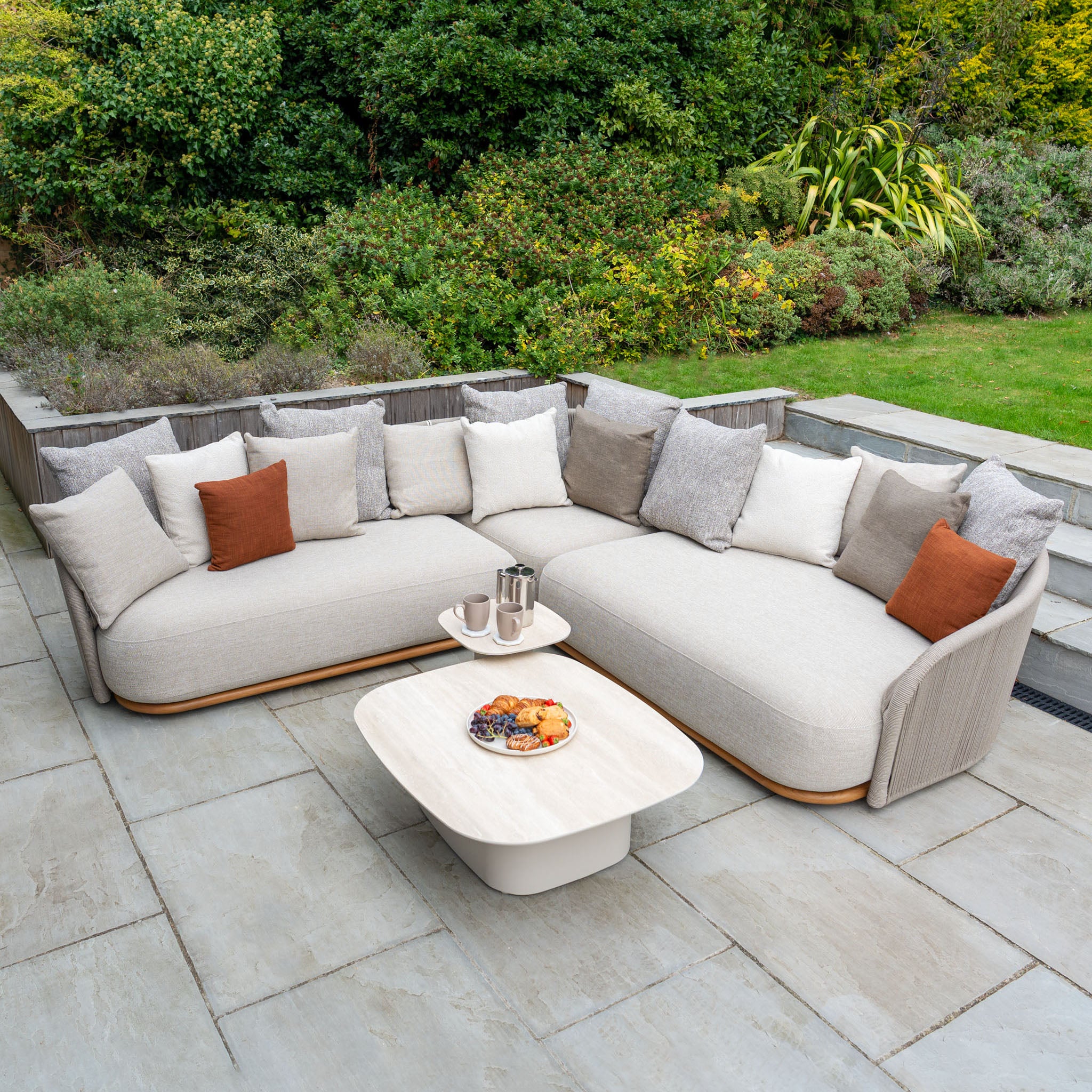 Avola Deluxe Corner Sofa Set in Fawn (Left Hand) with pillows, coffee table, and snacks on a stone patio—crafted from weather-resistant Olefin rope, premium teak, and aluminium for lasting garden comfort.