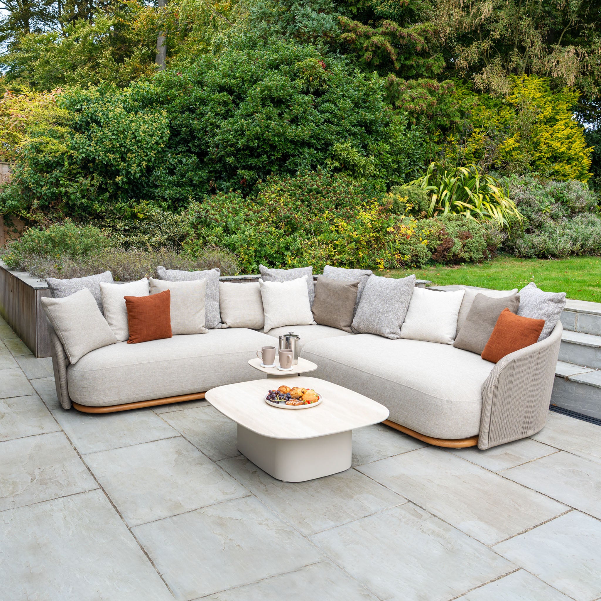 The Avola Deluxe Corner Sofa Set in Fawn (Left Hand) features plush cushions and a modern coffee table on a stone patio, combining weather-resistant design with teak and aluminium for stylish garden lounging.