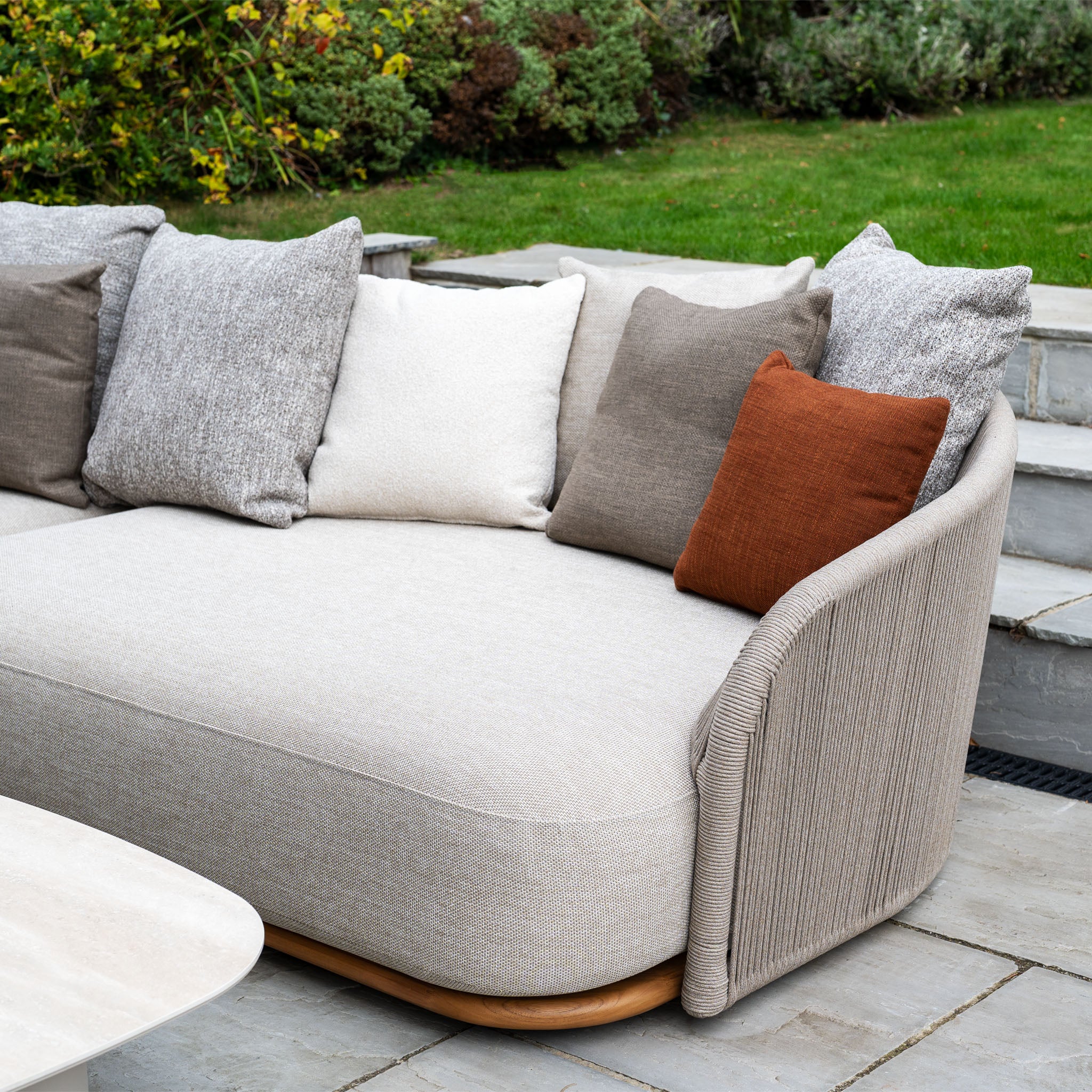 Avola Deluxe Corner Sofa Set in Fawn (Left Hand) with neutral and rust pillows, teak and aluminium garden details, and weather-resistant Olefin rope sits on a stone patio beside a green lawn.