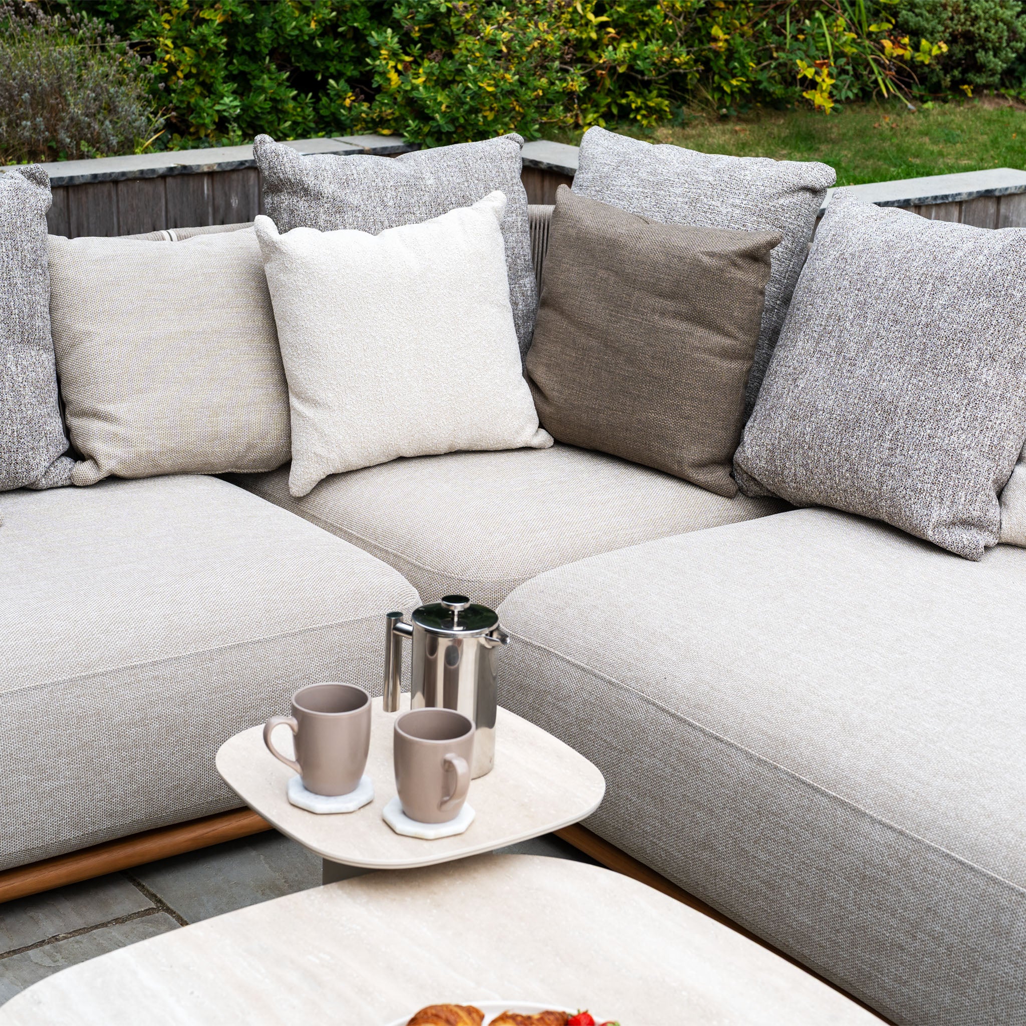The Avola Deluxe Corner Sofa Set in Fawn (Left Hand) features weather-resistant Olefin rope, assorted cushions, a coffee pot, and two mugs on a small table in a tranquil garden setting.