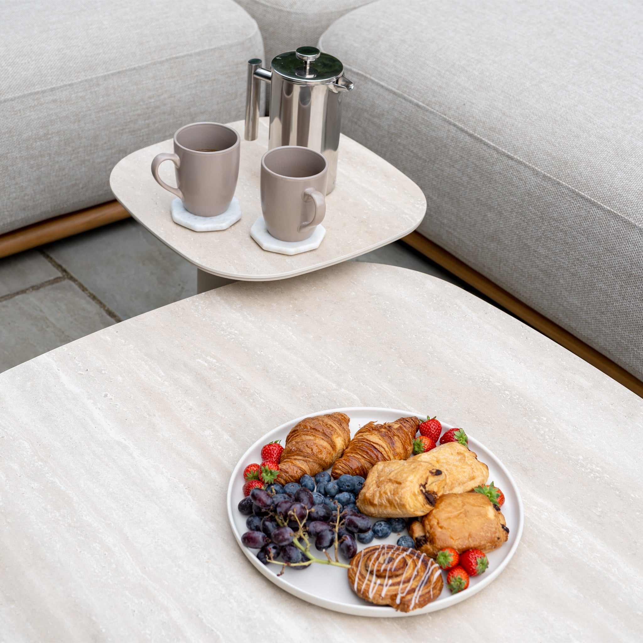 A plate of pastries, grapes, and strawberries sits next to two mugs and a coffee pot on a light table—perfect for sharing with friends while relaxing on your Avola Deluxe Corner Sofa Set in Fawn (Left Hand).