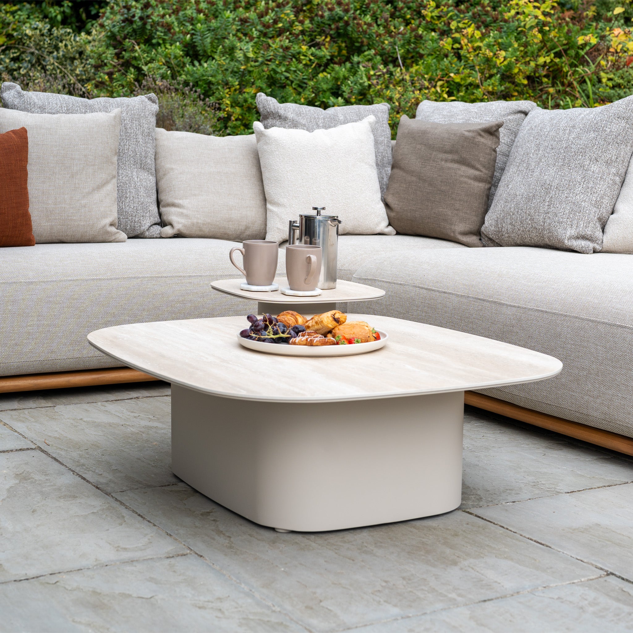 Modern outdoor seating area featuring teak and aluminium furniture, including the Avola Deluxe Corner Sofa Set in Fawn (Left Hand), with a low coffee table holding snacks, two cups, and a French press.