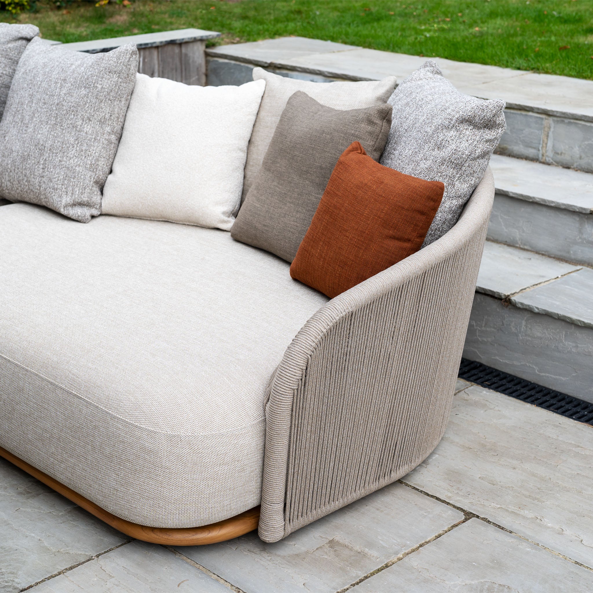 Avola Deluxe Corner Sofa Set in Fawn (Left Hand) with weather-resistant Olefin rope and teak-aluminium frame, styled with neutral and orange cushions on a stone patio by steps and grass.