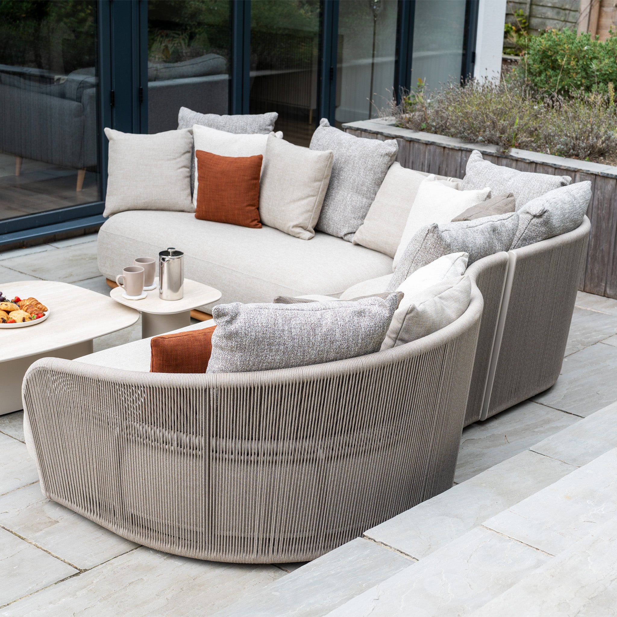 The Avola Deluxe Corner Sofa Set in Fawn (Left Hand) features beige cushions and a rope-wrapped base with weather-resistant Olefin rope, blending durable teak and aluminium elegance—ideal for patios.