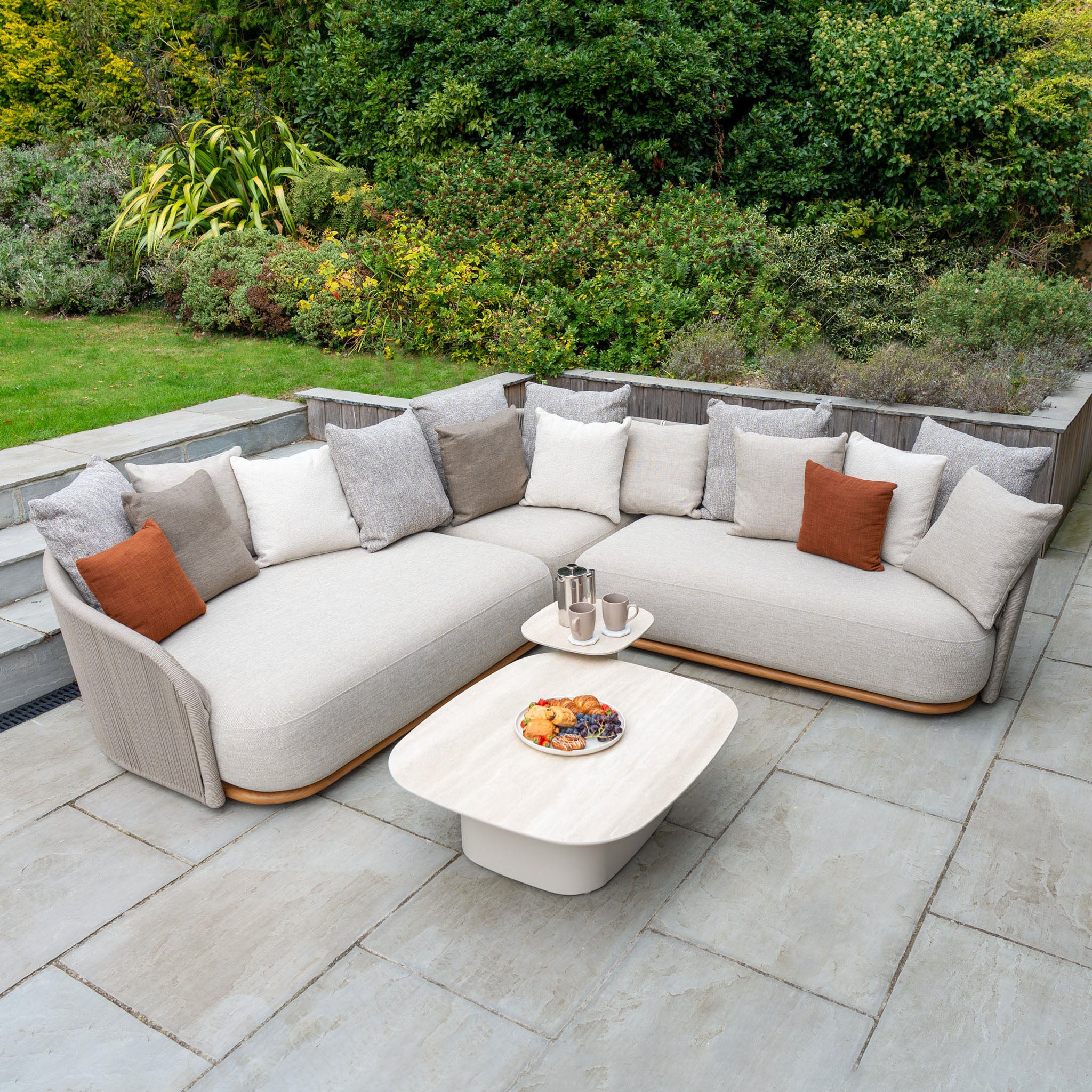 Modern outdoor patio with an Avola Deluxe Corner Sofa Set in Fawn (Right Hand) featuring weather-resistant Olefin rope, cozy pillows, a coffee table, and lush greenery in the background.
