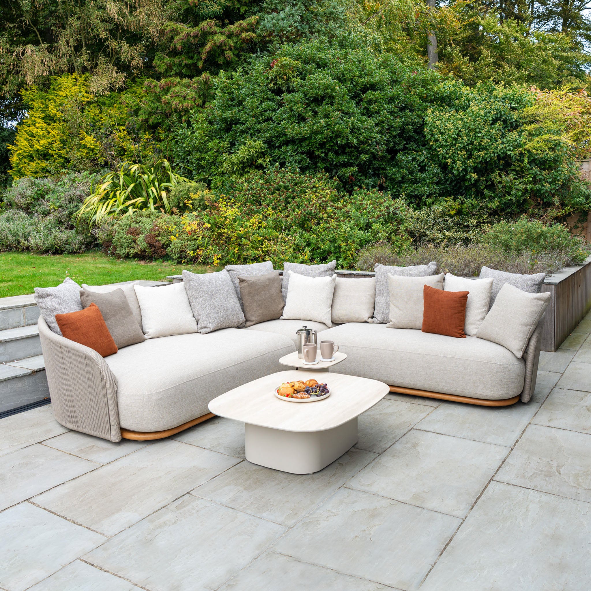 Avola Deluxe Corner Sofa Set in Fawn (Right Hand) with weather-resistant cushions and coffee table on a stone patio, surrounded by lush green bushes.
