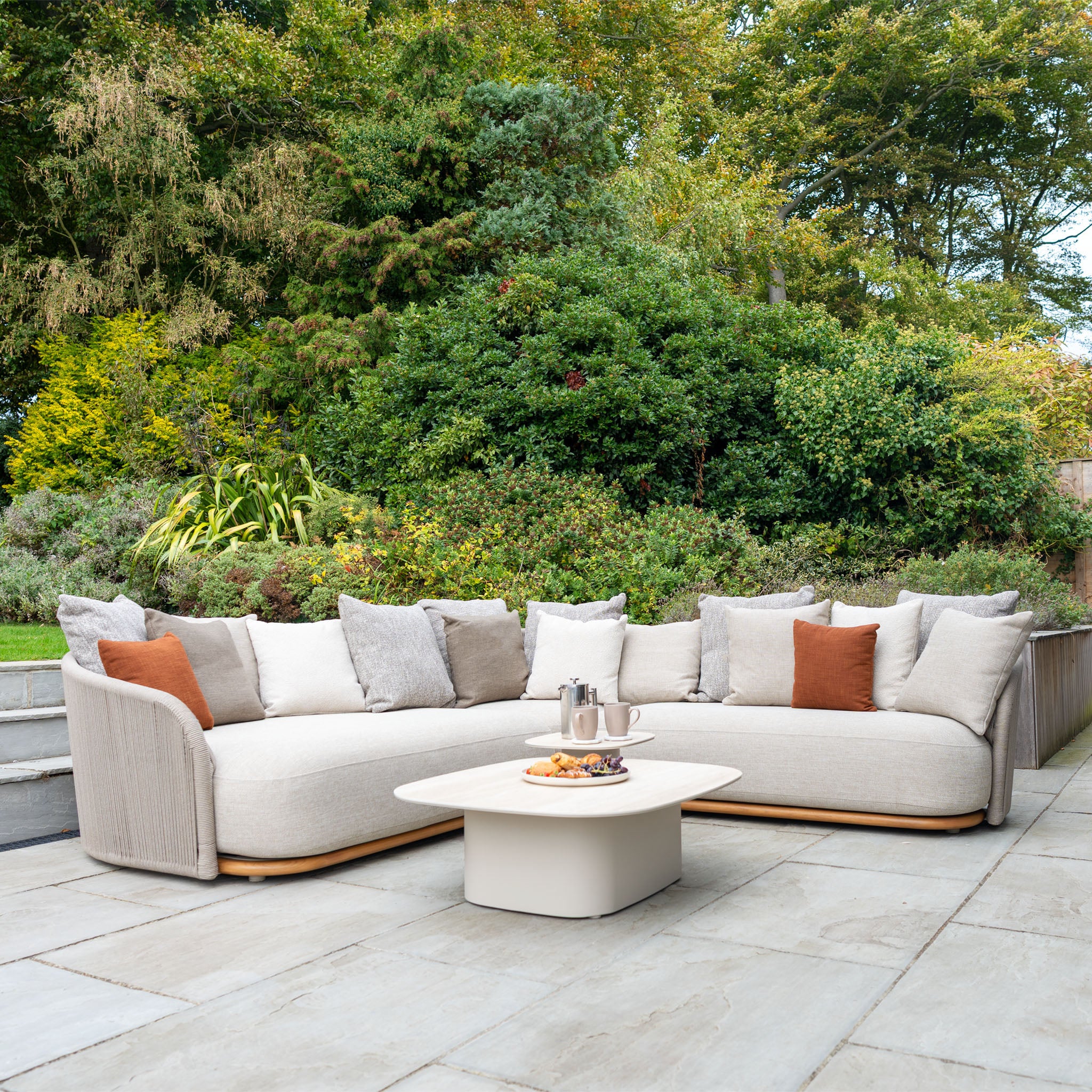 The Avola Deluxe Corner Sofa Set in Fawn (Right Hand) is a beige outdoor sectional with cushions and a coffee table, crafted from teak and aluminium, perfect for relaxing on a stone patio surrounded by greenery.
