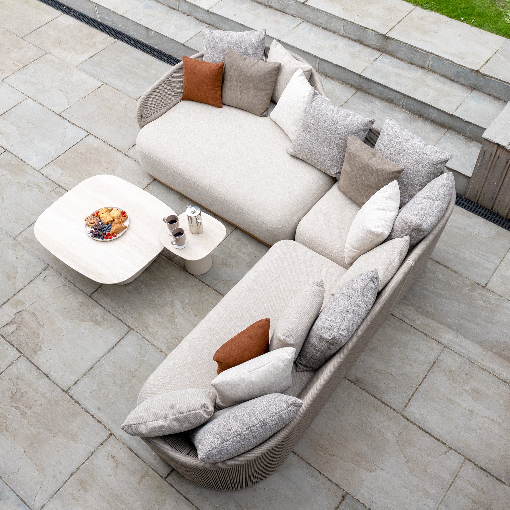 The Avola Deluxe Corner Sofa Set in Fawn (Right Hand) includes a modern teak and aluminium sofa with weather-resistant Olefin rope, plush cushions, a coffee table, coffee cups, and fruit plate arranged on a stone patio.