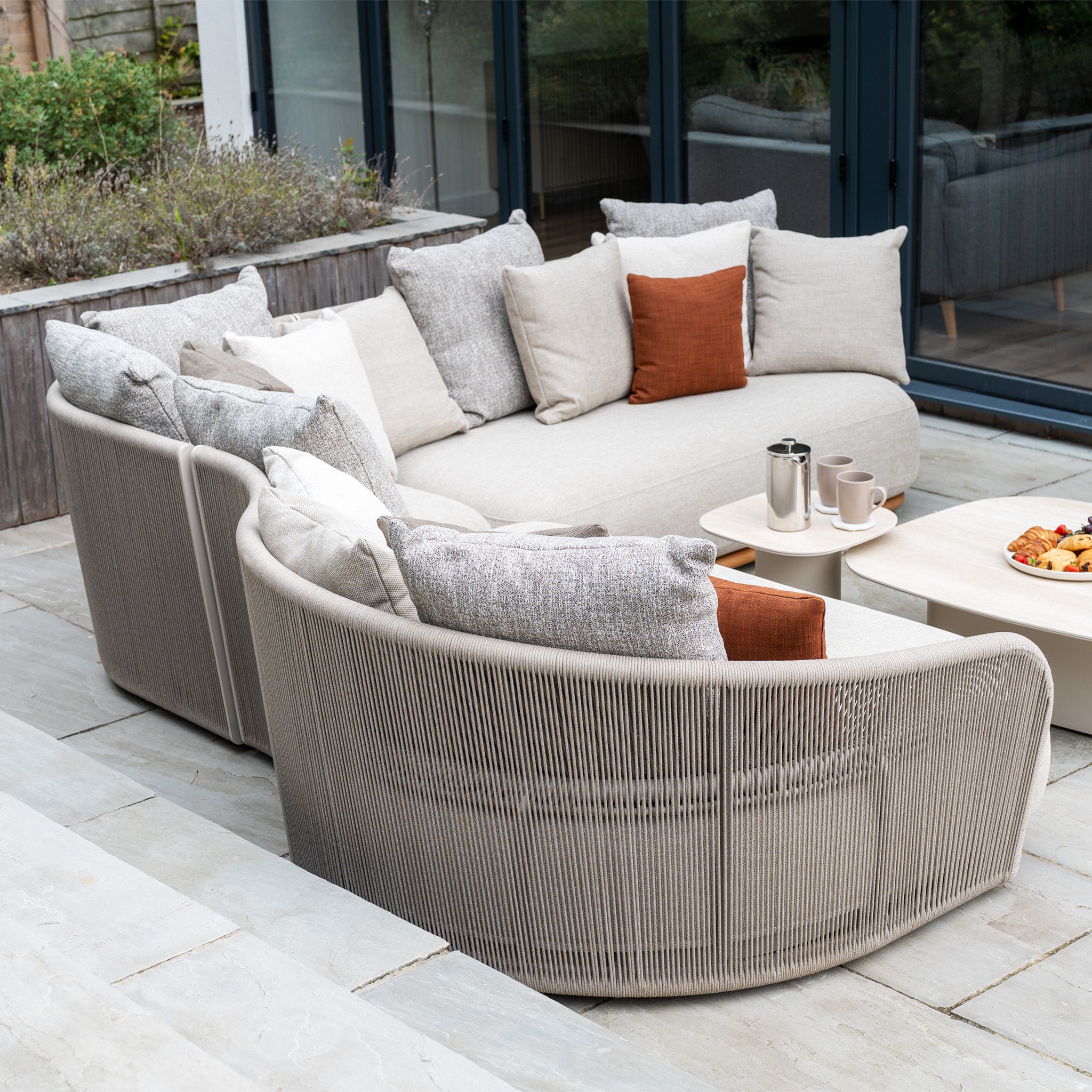 Modern outdoor patio set showcasing the Avola Deluxe Corner Sofa Set in Fawn (Right Hand) with beige weather-resistant sofas, gray and orange pillows, coffee mugs, and a tray of snacks on the table.
