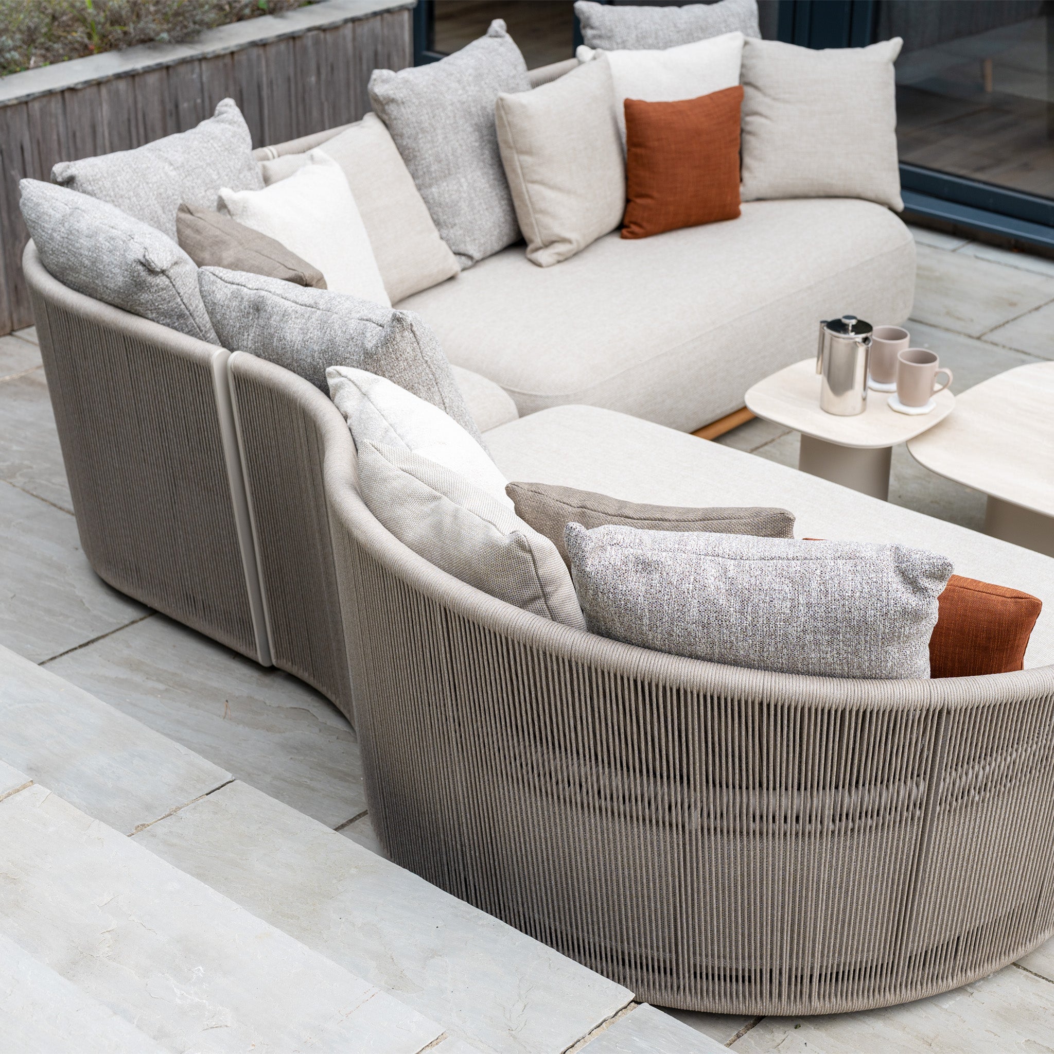 The Avola Deluxe Corner Sofa Set in Fawn (Right Hand) features neutral and rust-colored pillows on a stone patio beside a low table with mugs. Made from teak and aluminum, it has weather-resistant Olefin rope for lasting outdoor comfort.