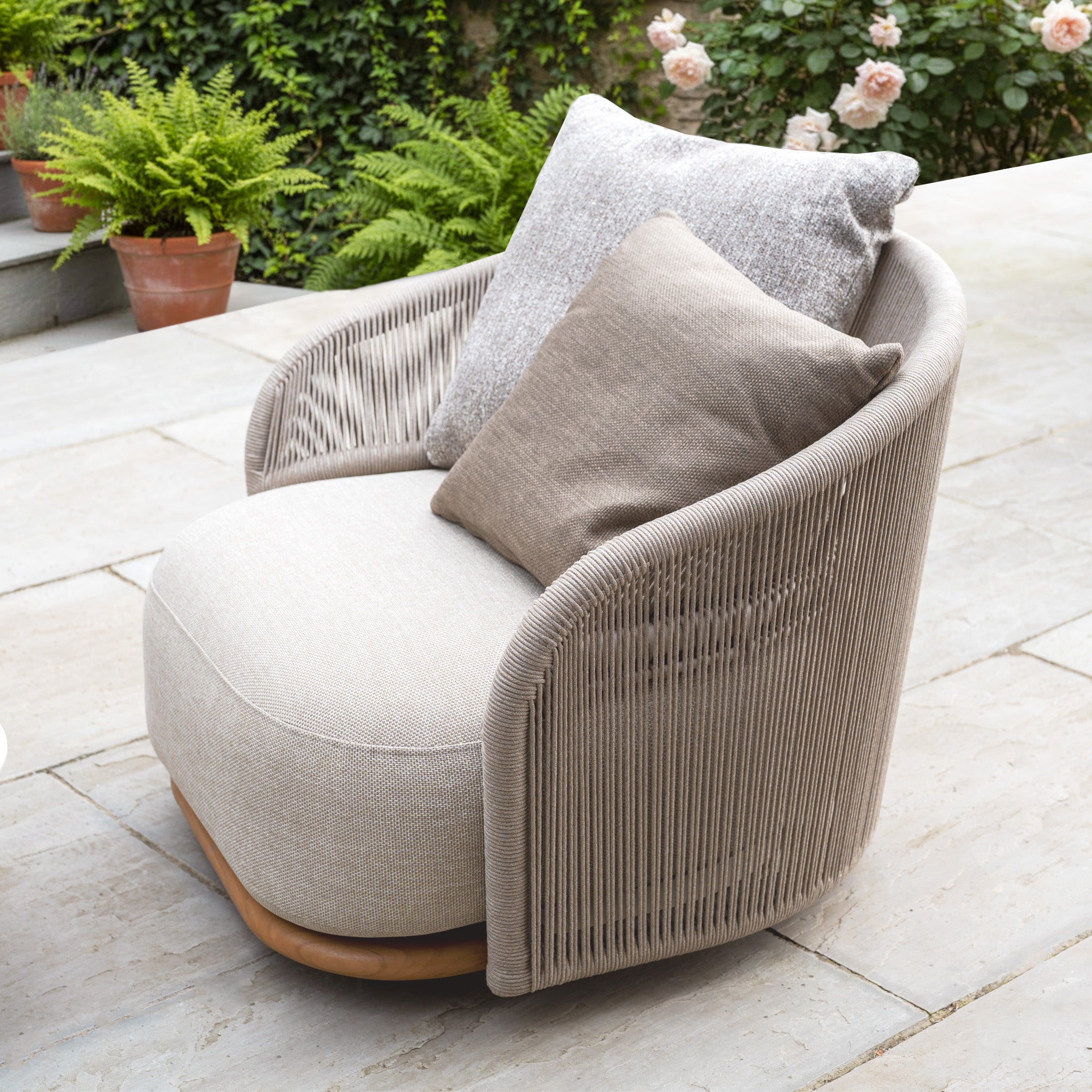 Avola Swivel Armchair in Fawn with two weather-resistant Olefin cushions is shown on a stone patio beside potted plants and flowers, making it ideal for outdoor living.