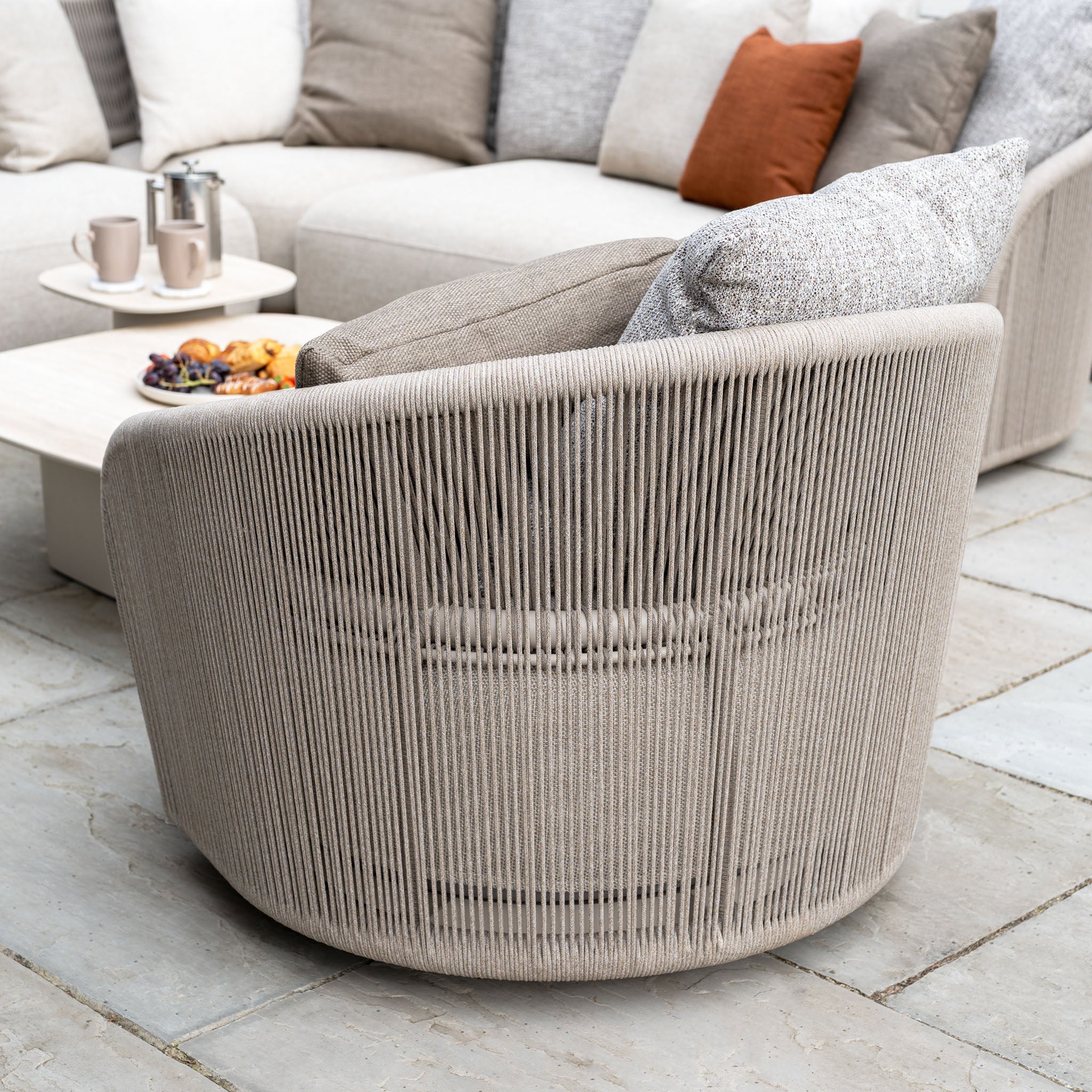 The Avola Swivel Armchair in Fawn with weather-resistant Olefin cushions sits on a stone patio, next to a coffee table with snacks and cups.