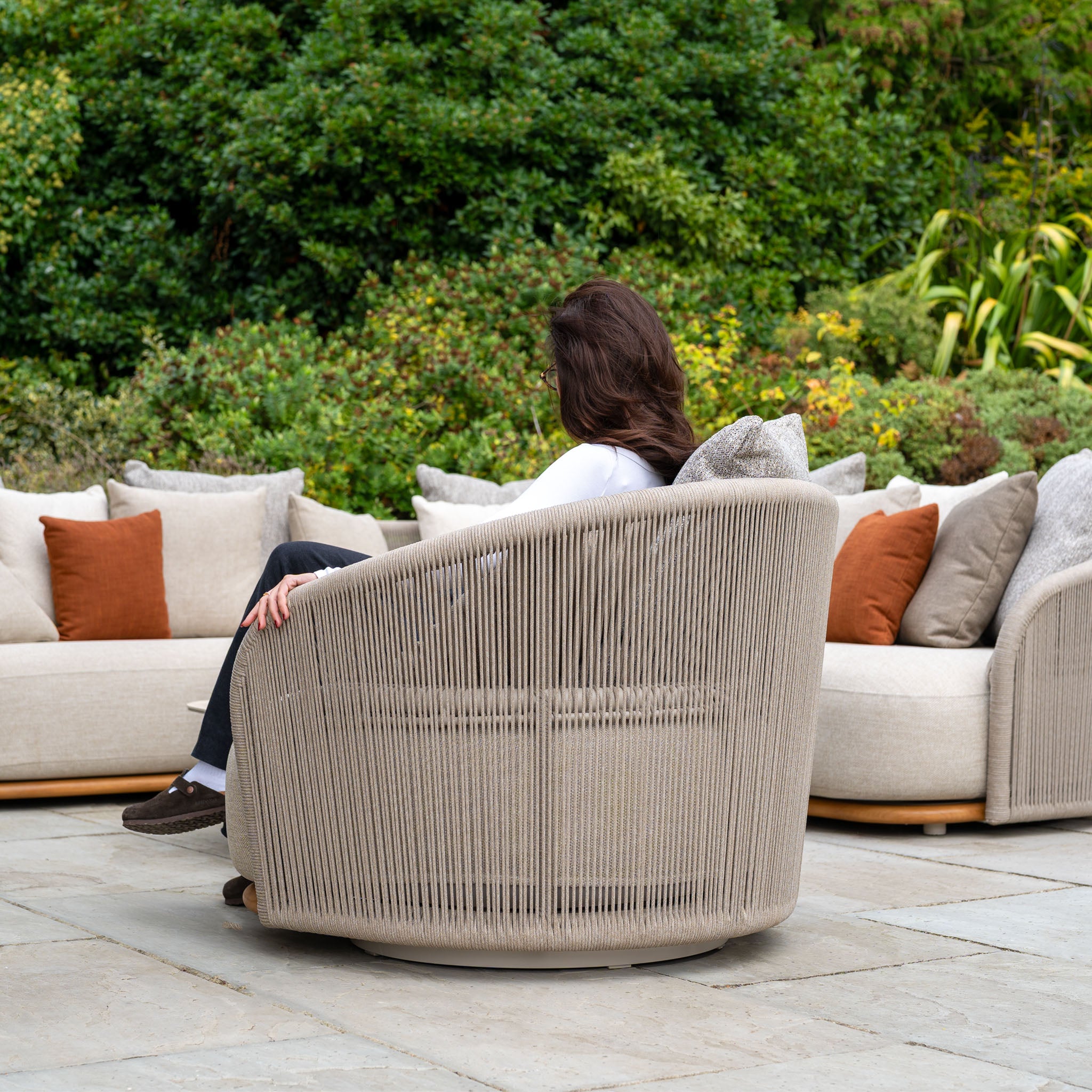 Person seated on an Avola Swivel Armchair in Fawn, facing away amid cushioned sofas and lush greenery. Weather-resistant Olefin upholstery makes this outdoor chair ideal for relaxing outdoors year-round.