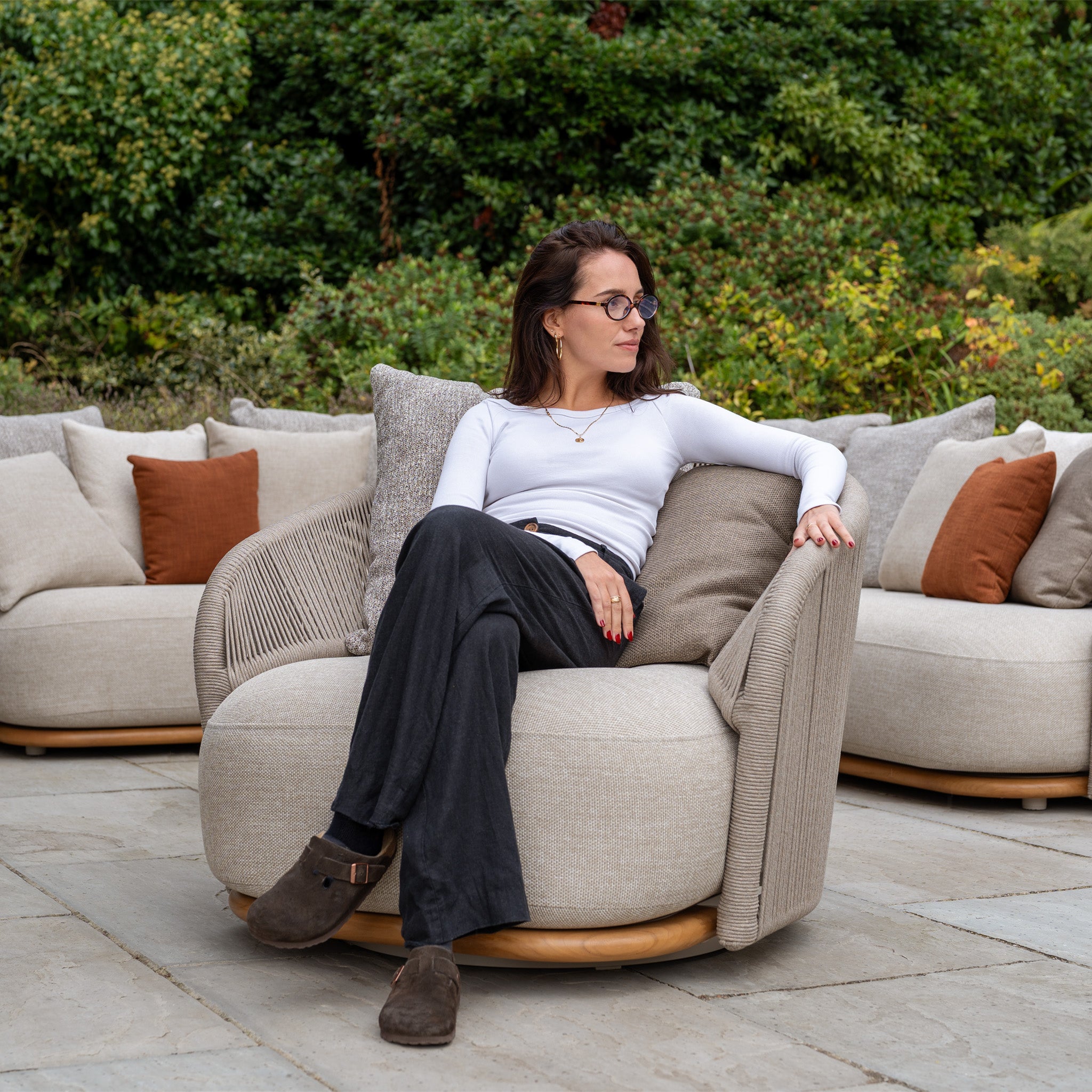 A woman with glasses sits on a beige Avola Swivel Armchair in Fawn, an outdoor chair made with weather-resistant Olefin, surrounded by sofas and lush greenery.