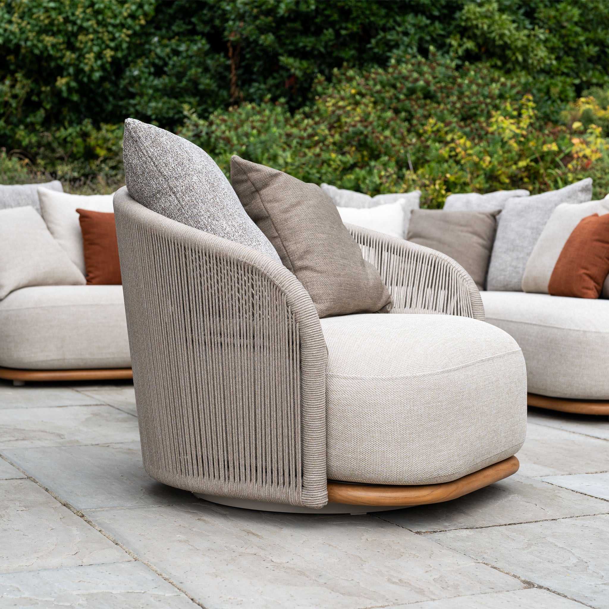 Avola Swivel Armchair in Fawn with weather-resistant Olefin cushions on a stone patio, surrounded by a sectional sofa and greenery—ideal for relaxing in your outdoor living setup.