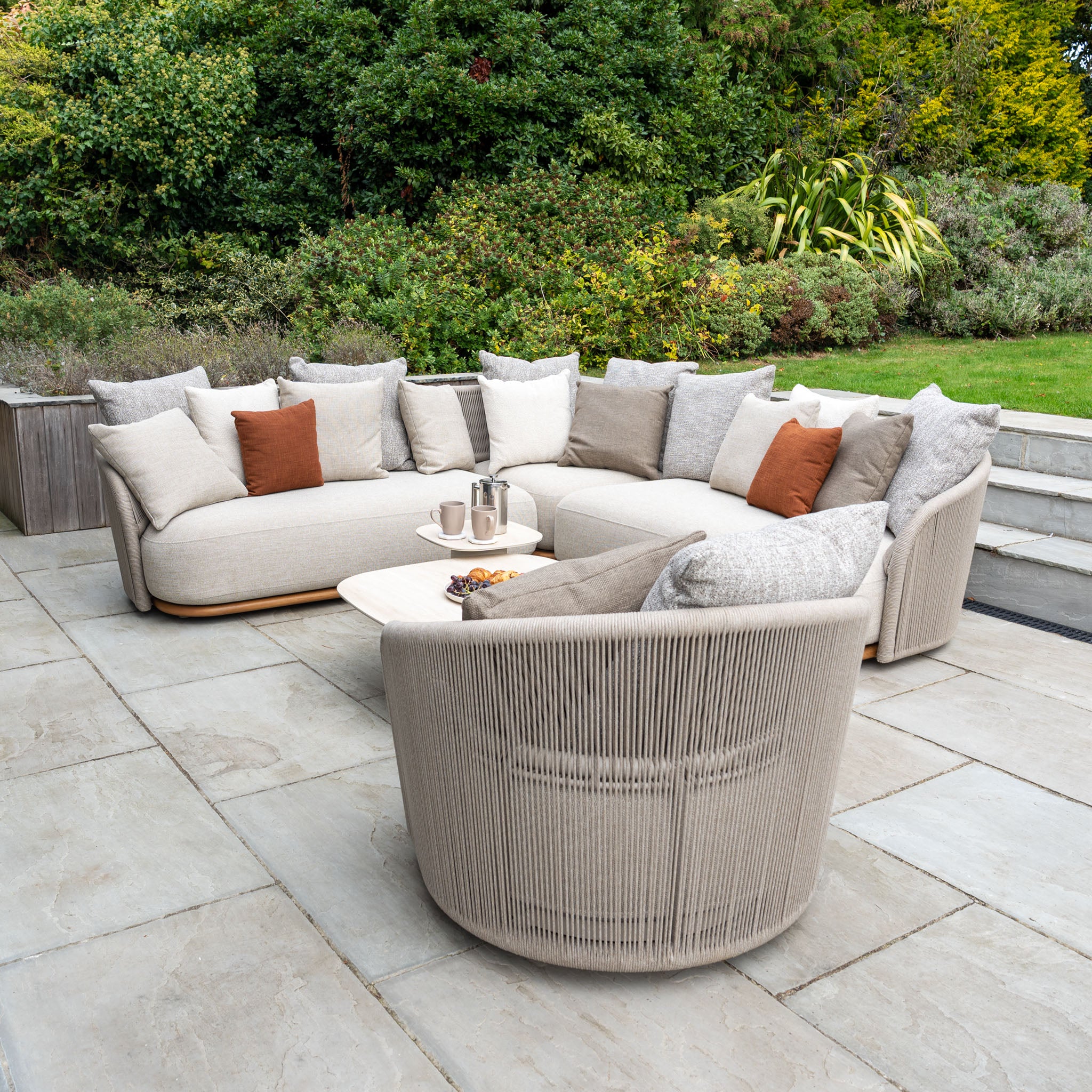 Modern outdoor patio with beige cushioned furniture, featuring the Avola Swivel Armchair in Fawn crafted with weather-resistant Olefin, a coffee table, and greenery in the background.