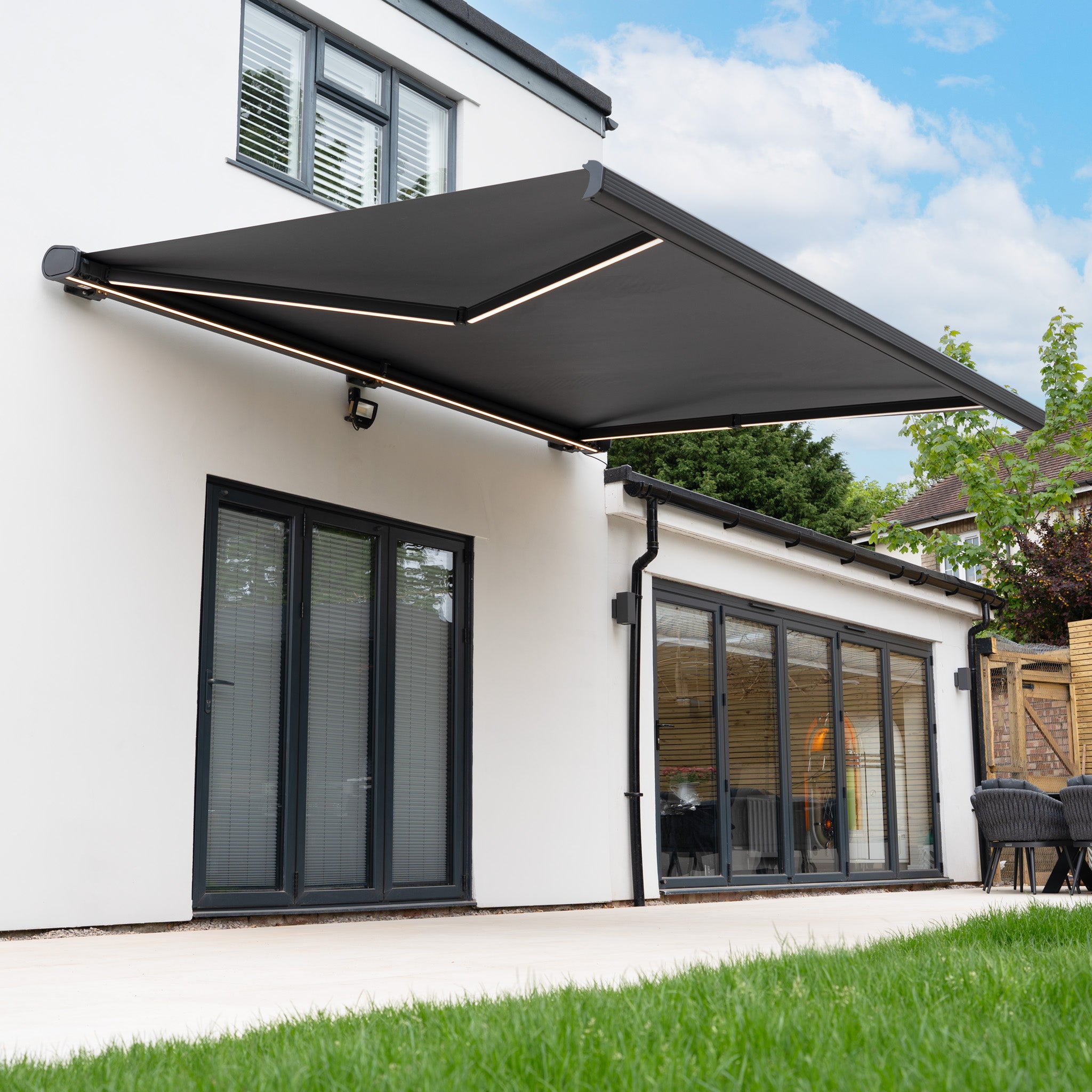 A modern house features a 3m x 2m Electric Motorised Awning in Grey with waterproof acrylic fabric, fully extended over the patio and glass doors on a sunny day.