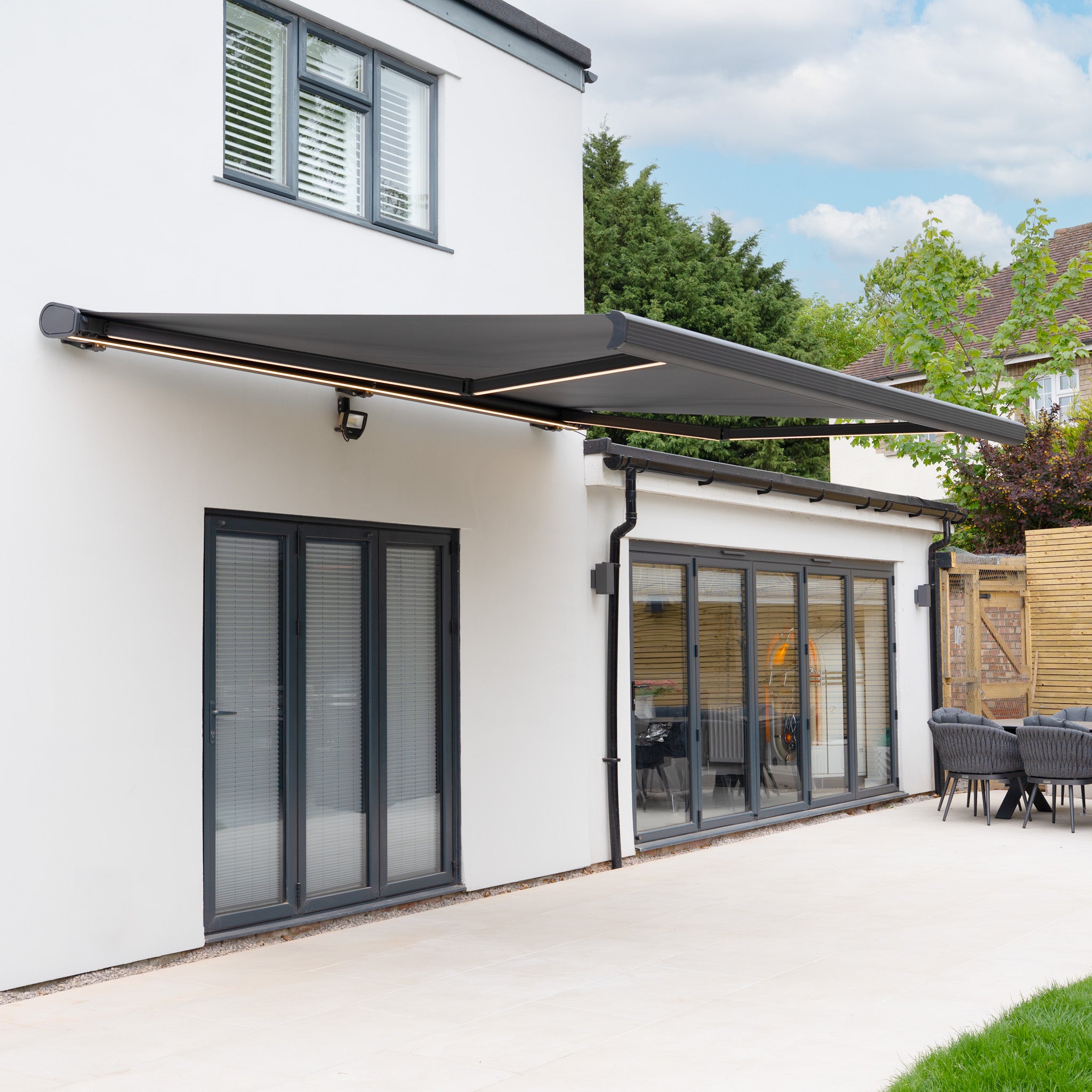 A modern house with large glass doors features a 3m x 2m Electric Motorised Awning in Grey, made from waterproof acrylic fabric, extended over the patio area.