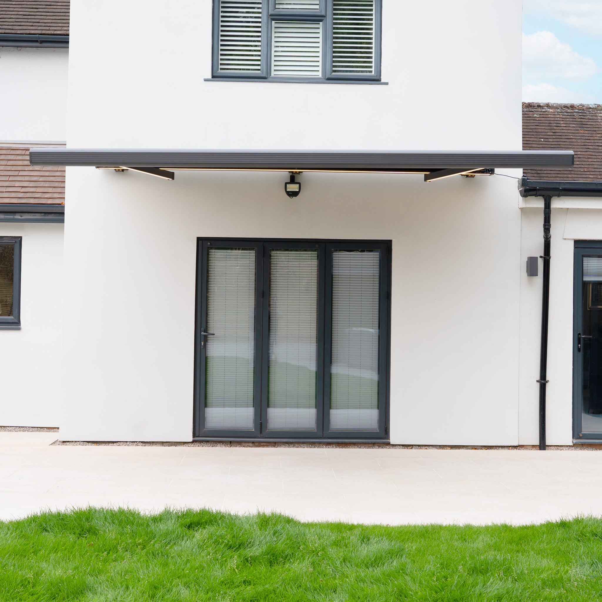 Modern house exterior with glass doors, a 3m x 2m Electric Motorised Awning in Grey featuring waterproof acrylic fabric, and a green lawn in the foreground.