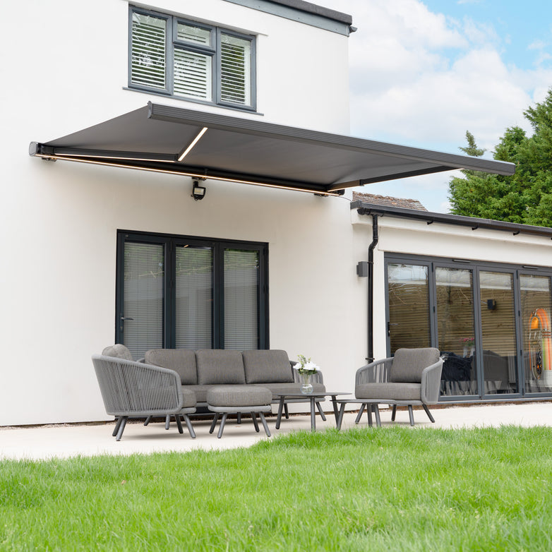 A modern patio with grey outdoor furniture sits beneath a 4m x 3m Electric Motorised Awning in Grey, creating a stylish outdoor living space attached to a white house.