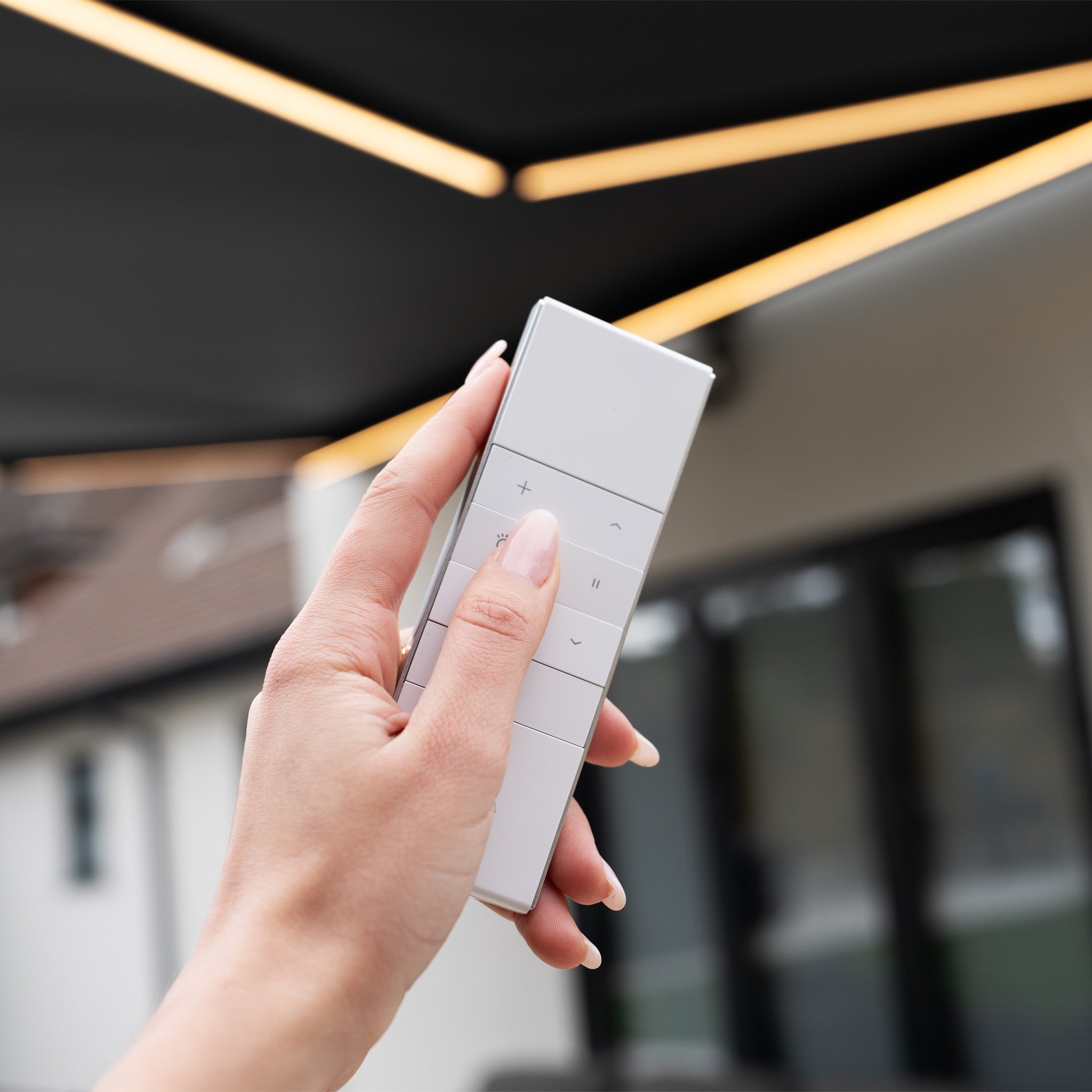 A hand holds a white remote in front of a modern building, operating the 3m x 2m Electric Motorised Awning in Grey made from waterproof acrylic fabric.