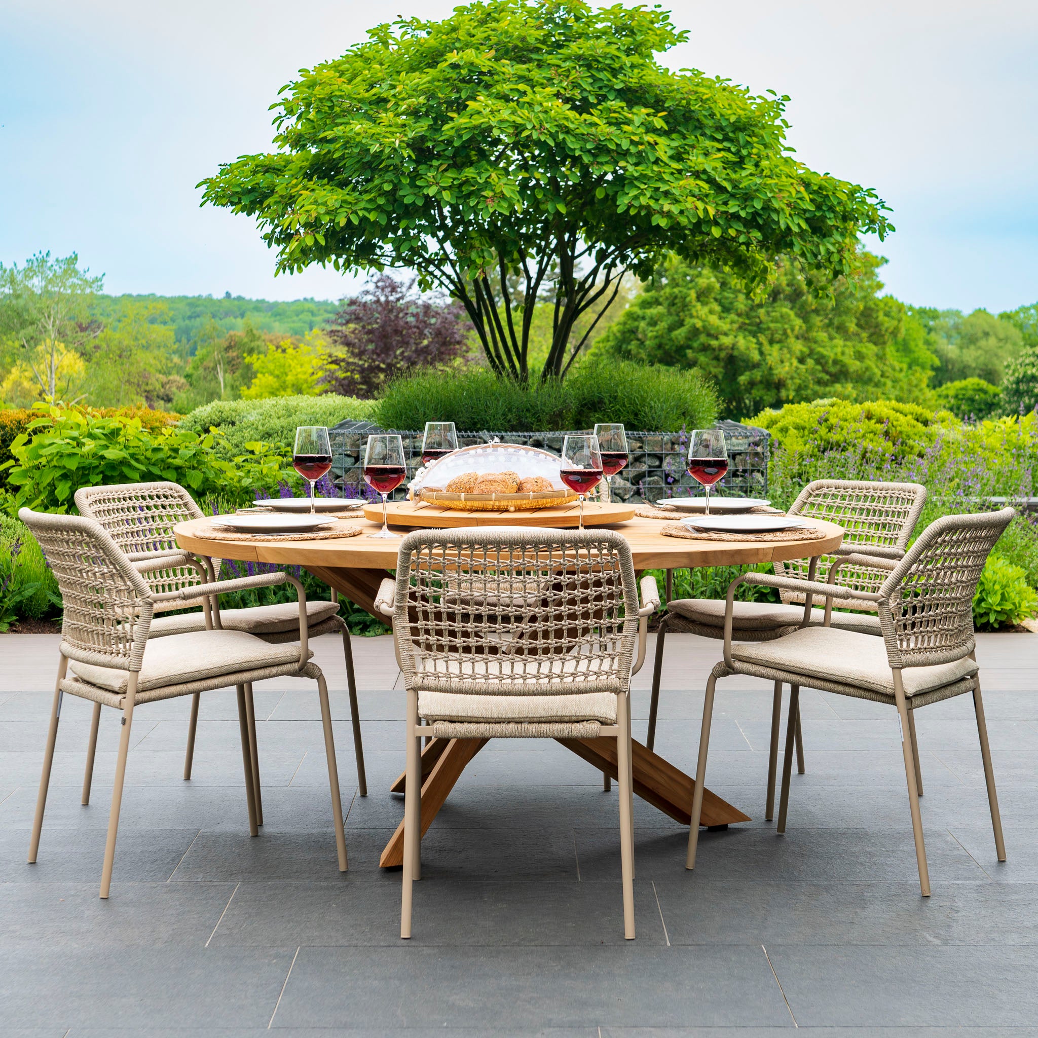 The Barista 6 Seat Round Dining Set with Lazy Susan in Latte is beautifully adorned with wine glasses and plates, encircled by a lush green landscape. This 4 Seasons outdoor setup, crafted with polyethylene core ropes for enhanced durability and style, is perfect for any occasion.