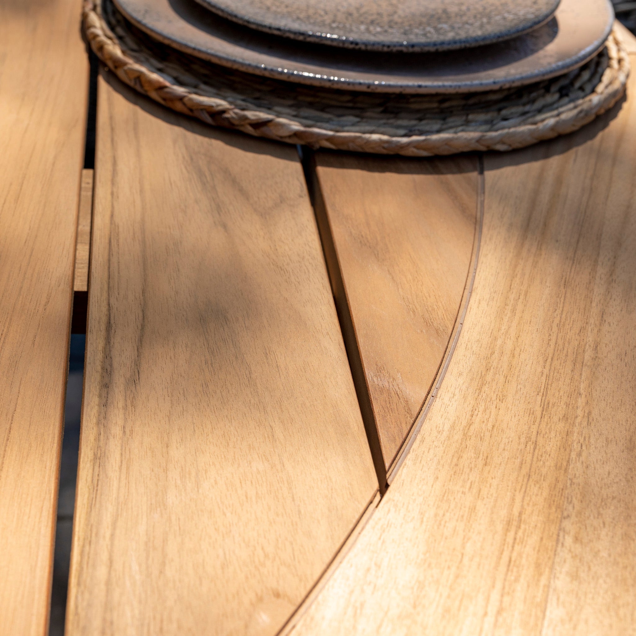 Close-up of a teak wood table from the Puccini 6 Seat Round Dining Set with Lazy Susan in Latte, featuring a decorative woven placemat and ceramic plates in a natural sunlight setting.