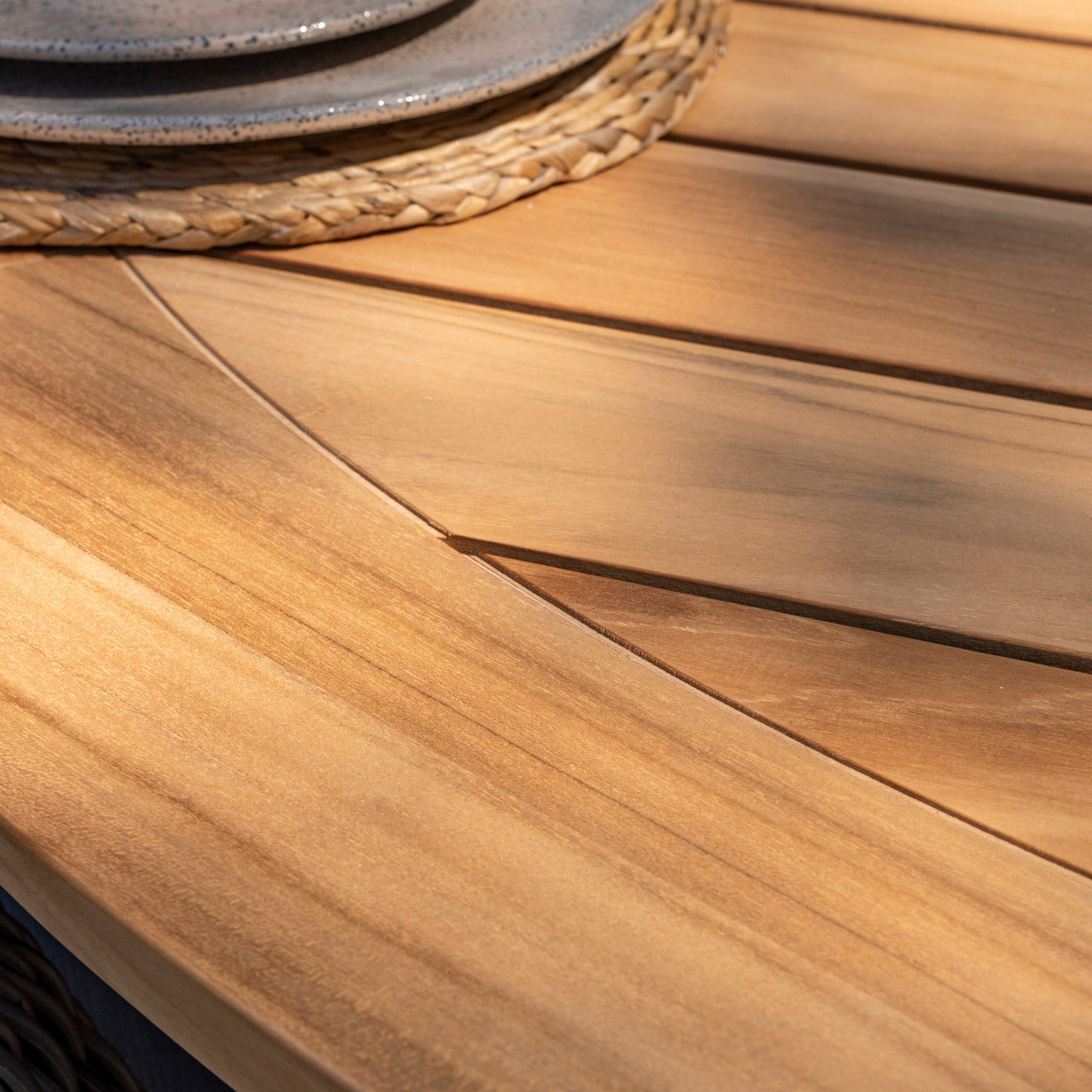 Close-up of a wooden table with neatly arranged plates on a woven mat. Sunlight casts a shadow on the smooth surface, highlighting the elegance perfect for outdoor dining in any season with the Barista 6 Seat Round Dining Set with Lazy Susan in Latte complementing the setting.