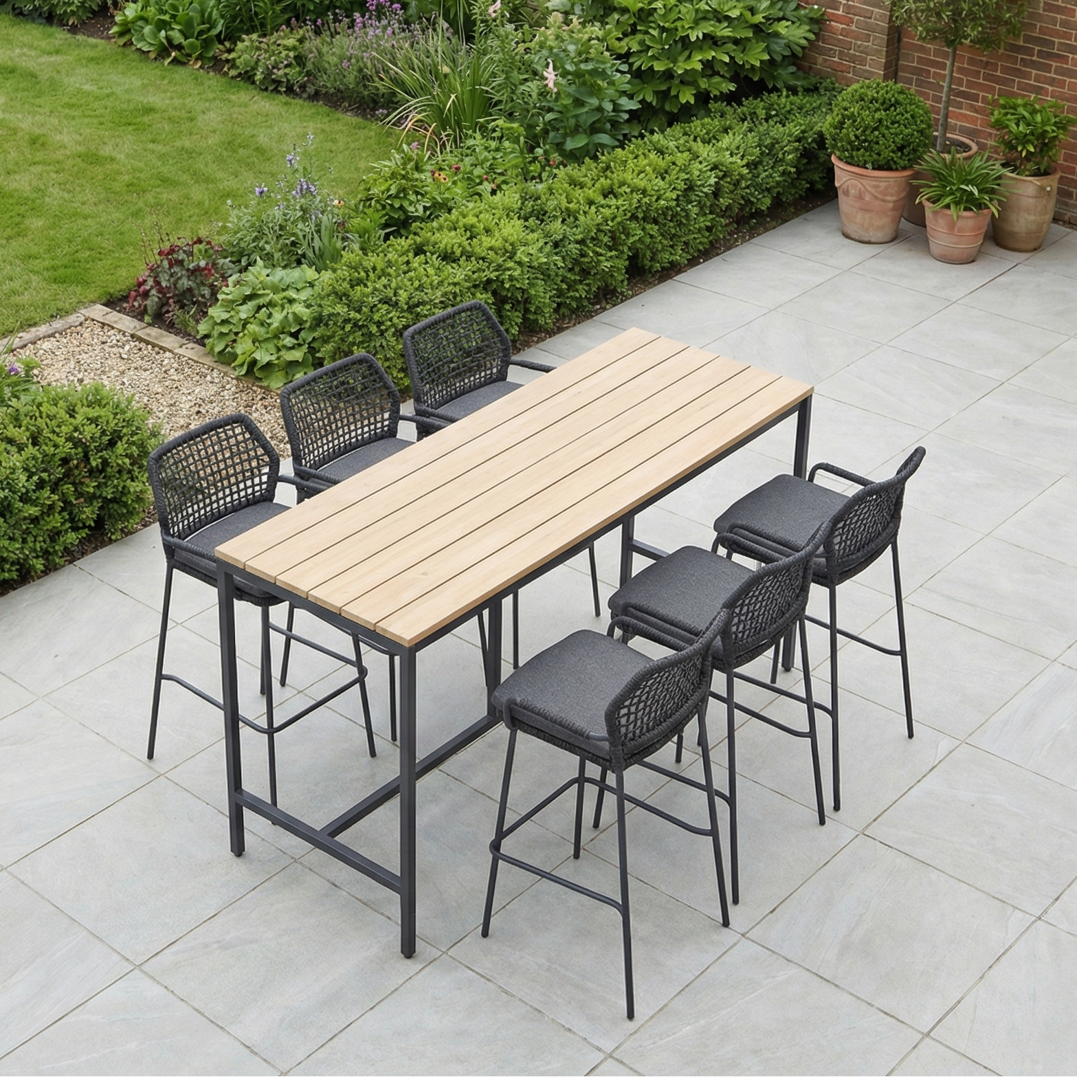 The Barista 6 Seat Bar Set with Teak Table in Anthracite features a teak table and six black chairs, perfect for relaxing outdoors on a tiled patio surrounded by lush greenery.