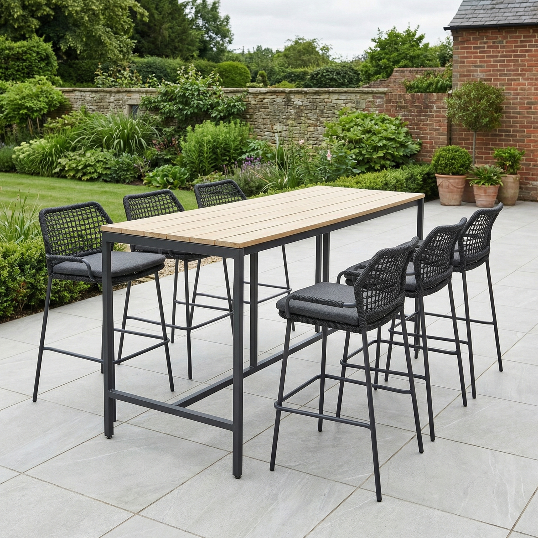 Enjoy the Barista 6 Seat Bar Set with Teak Table in Anthracite—tall table and six black chairs on tiled patio, surrounded by greenery. Perfect for stylish outdoor gatherings.