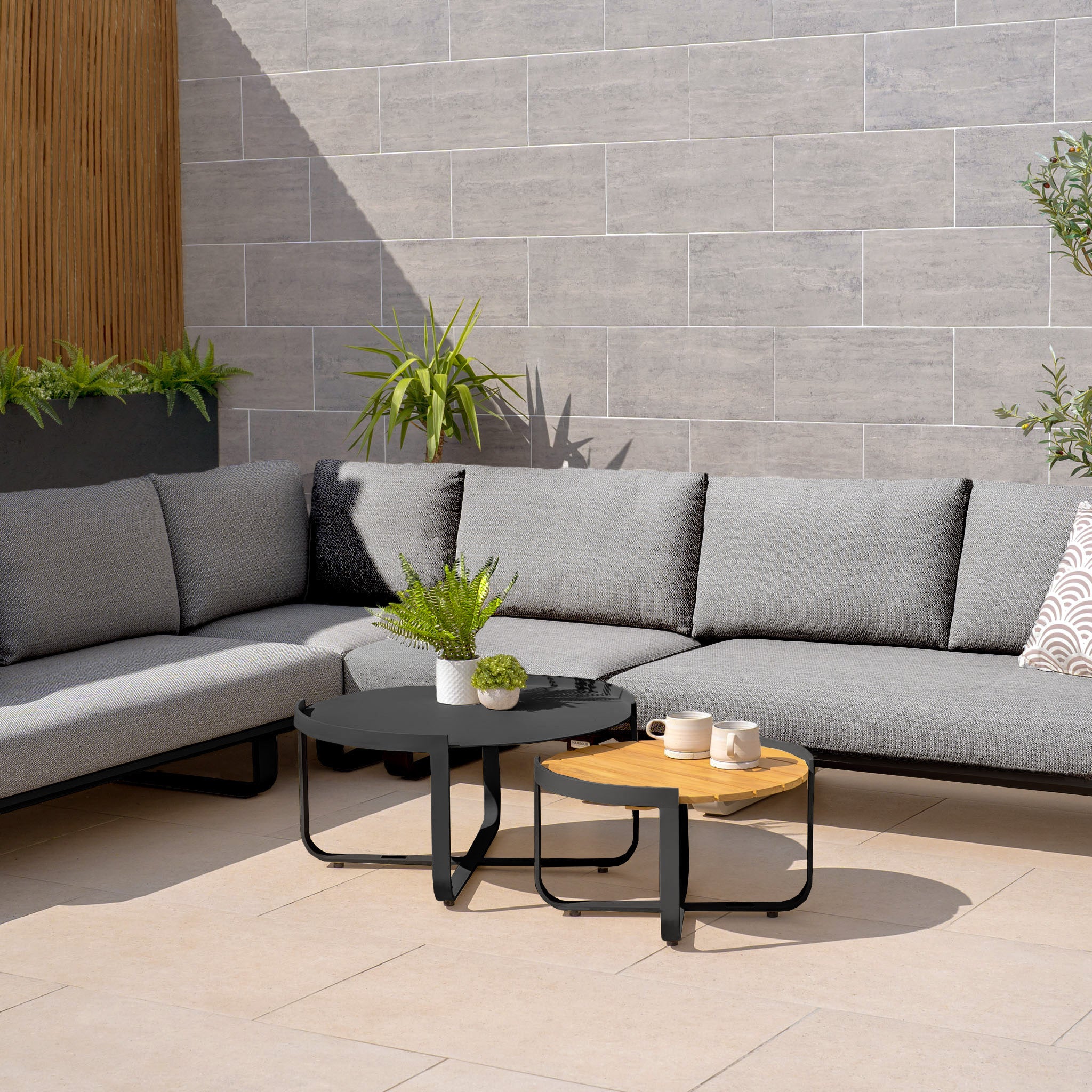 Modern outdoor patio featuring a Bay Large Corner Group Set with Aluminium Frame in Charcoal, including a gray sectional, two round coffee tables, and green plants against a tiled wall. The sofa is designed with durable Olefin material set on a powder-coated aluminum frame.