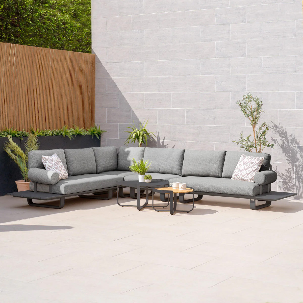 An outdoor patio showcases the Bay Large Corner Group Set with an aluminum frame in charcoal, paired with side tables and greenery against a light brick wall. The ambiance is further enhanced by an outdoor dining set, providing both functionality and style to this peaceful outdoor retreat.