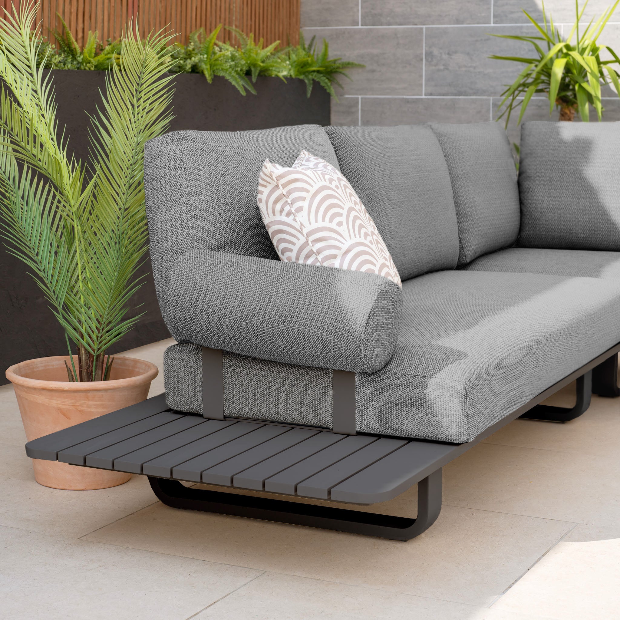The Bay Large Corner Group Set with Aluminium Frame in Charcoal, crafted from durable Olefin material and featuring a powder-coated aluminium frame, includes a gray outdoor sofa adorned with a patterned pillow, perfectly situated next to potted plants on a tiled patio.