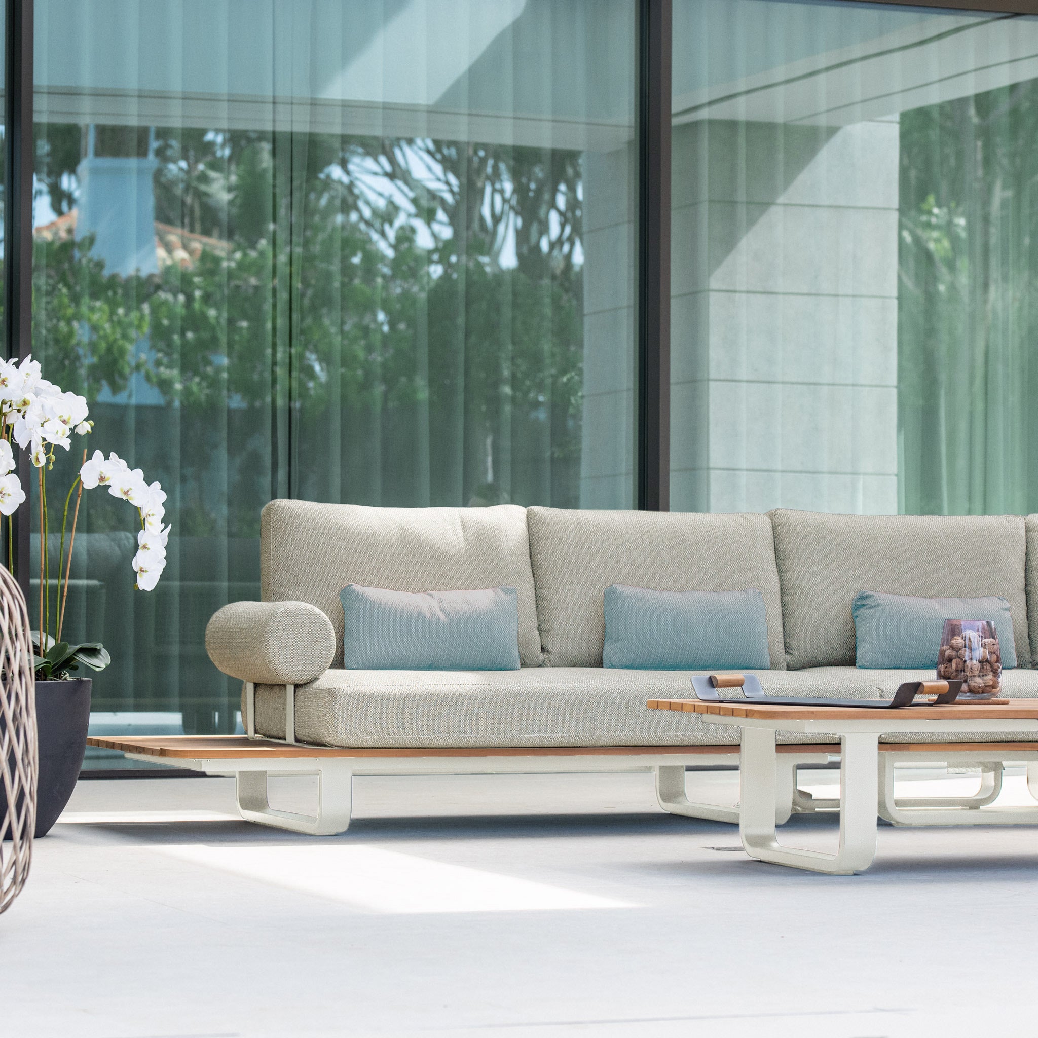 Modern outdoor seating area featuring the Bay Large Corner Group Set with 4 Waist Pillows and Teak Table in Latte, large glass windows, and potted orchid flowers in view.