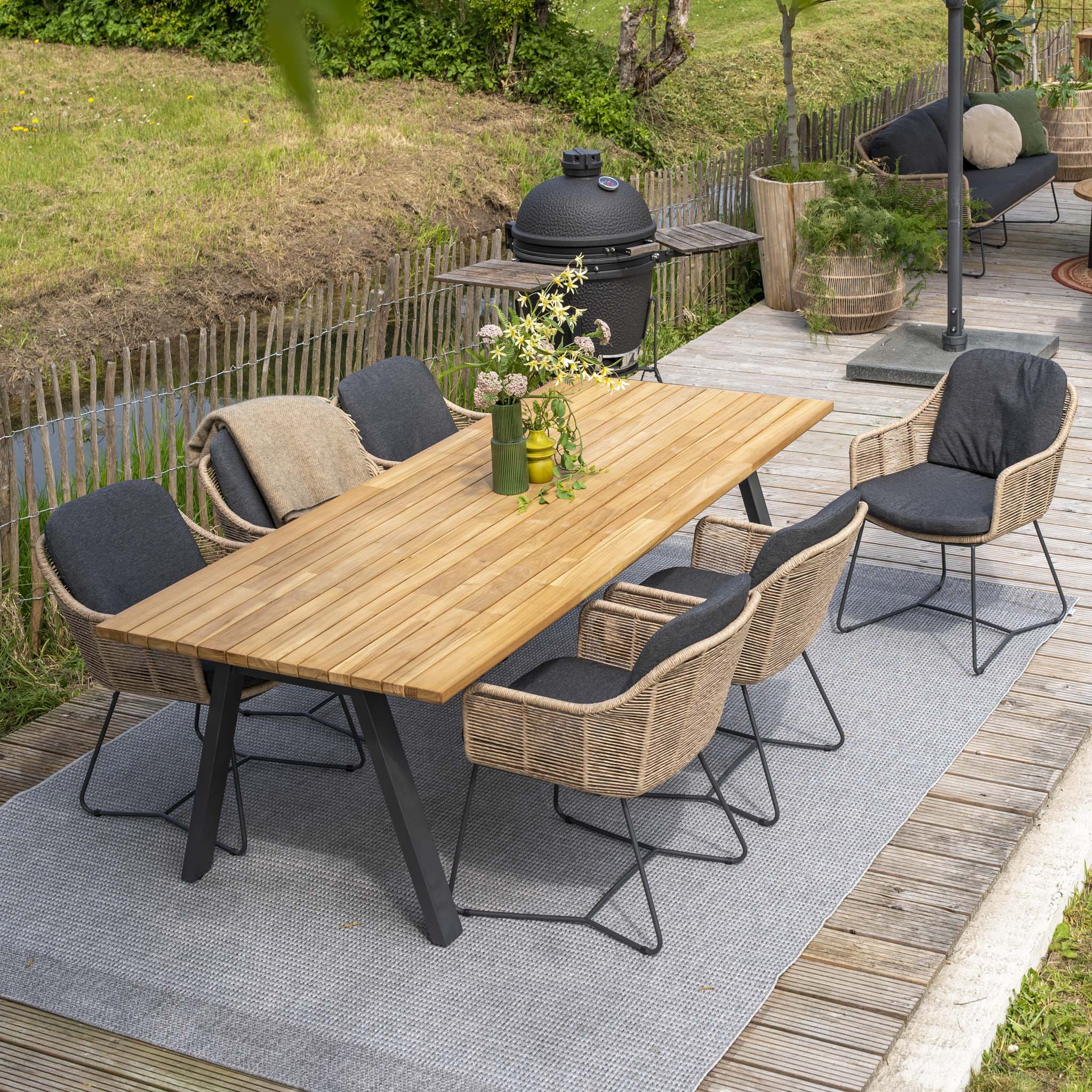 Outdoor dining area with the Belmond 6 Seat Dining Set featuring a teak table in natural finish, hand-woven natural poly rattan chairs, and a grill on a deck surrounded by greenery. The elegant wicker chairs enhance the serene setting perfectly.
