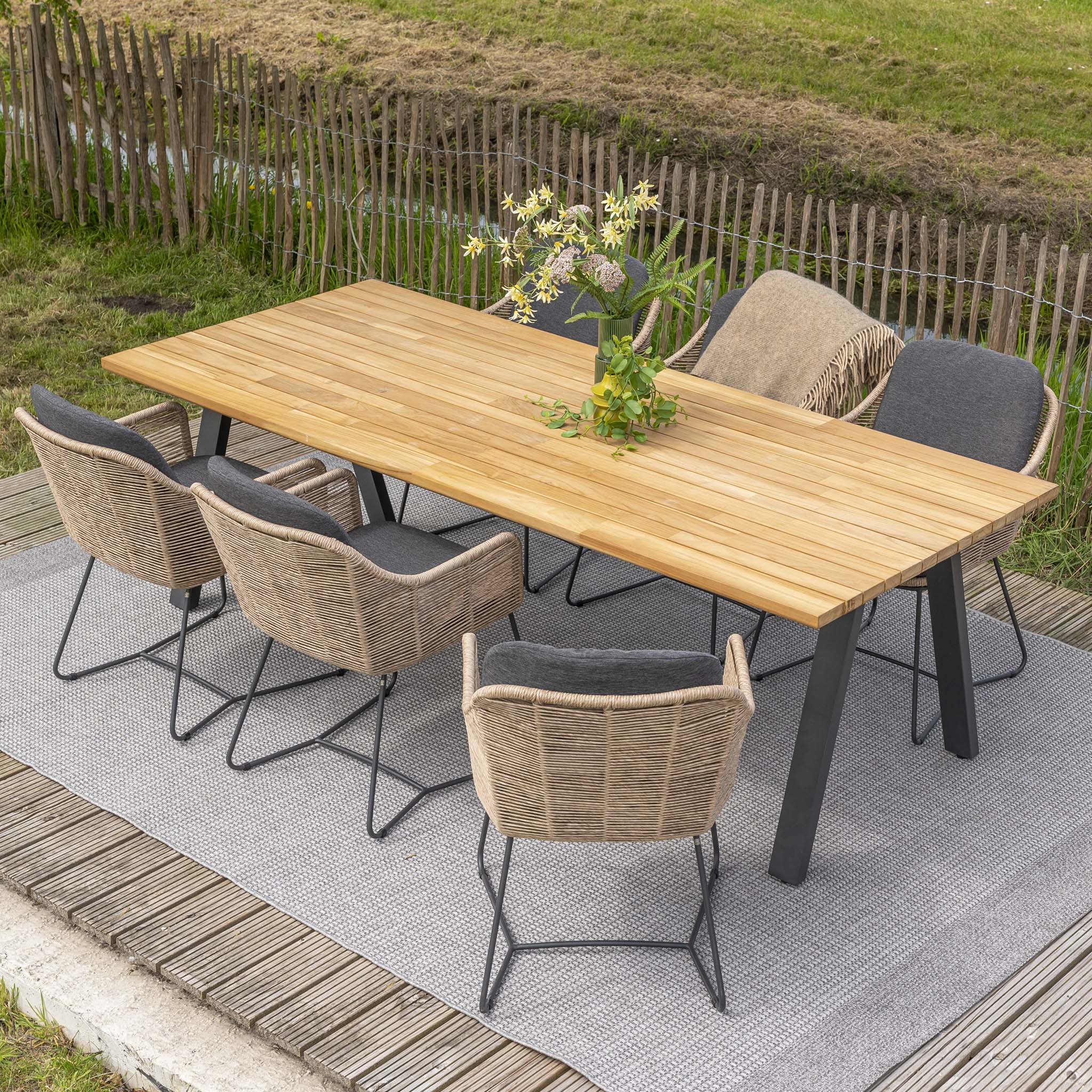 The Belmond 6 Seat Dining Set with Teak Table in Natural features a stunning outdoor wooden table paired with wicker chairs, all set on a gray rug. The hand-woven natural poly rattan chairs, each adorned with a blanket draped over them, offer both stylish and weatherproof seating for your patio. A vase filled with flowers completes this elegant Belmond range arrangement.
