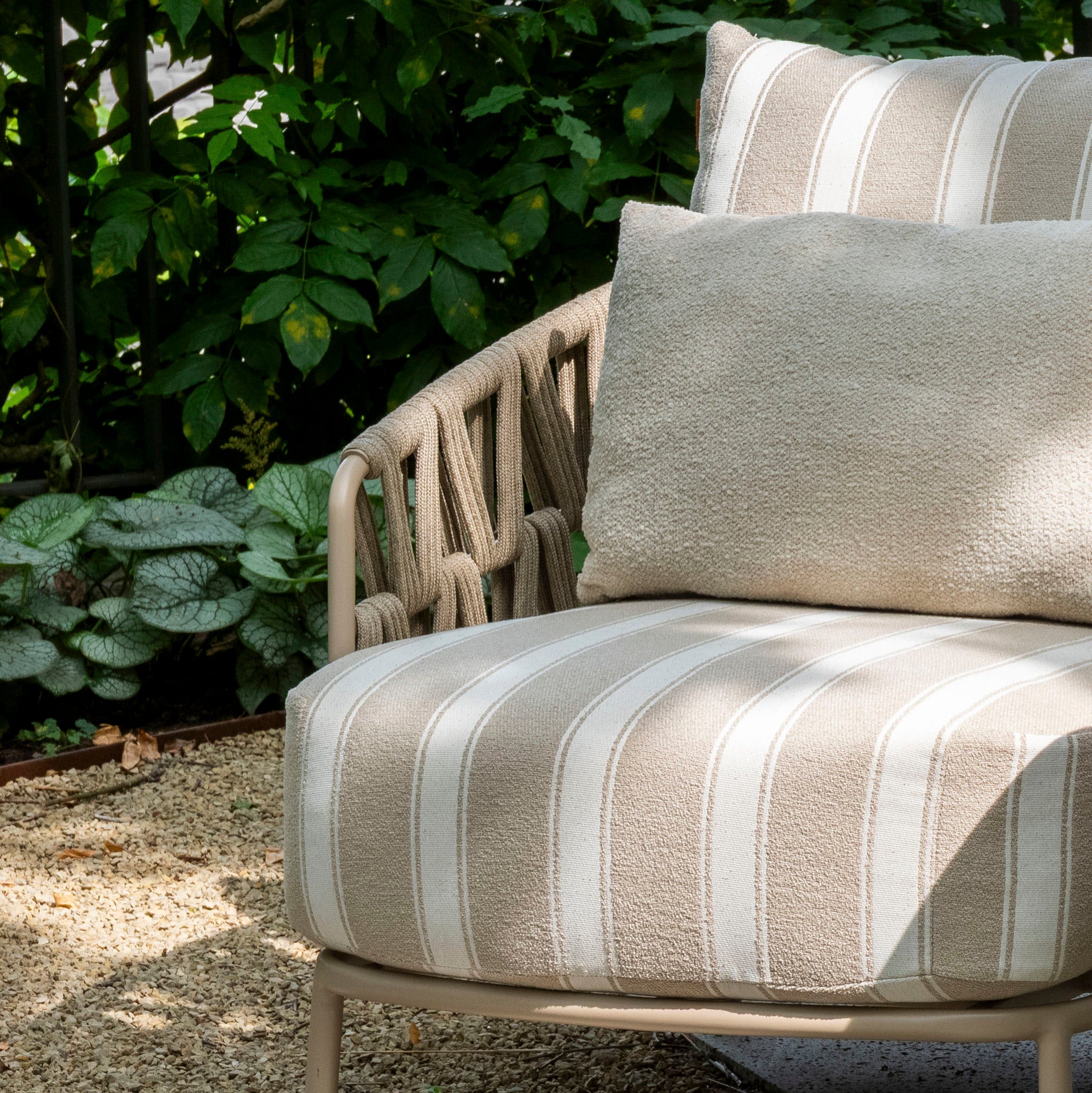 The Madeira 3 Seat Sofa Set in Striped Latte features a beige outdoor sofa with striped cushions and two pillows, creating a stylish garden furniture setup on gravel beside lush green plants.