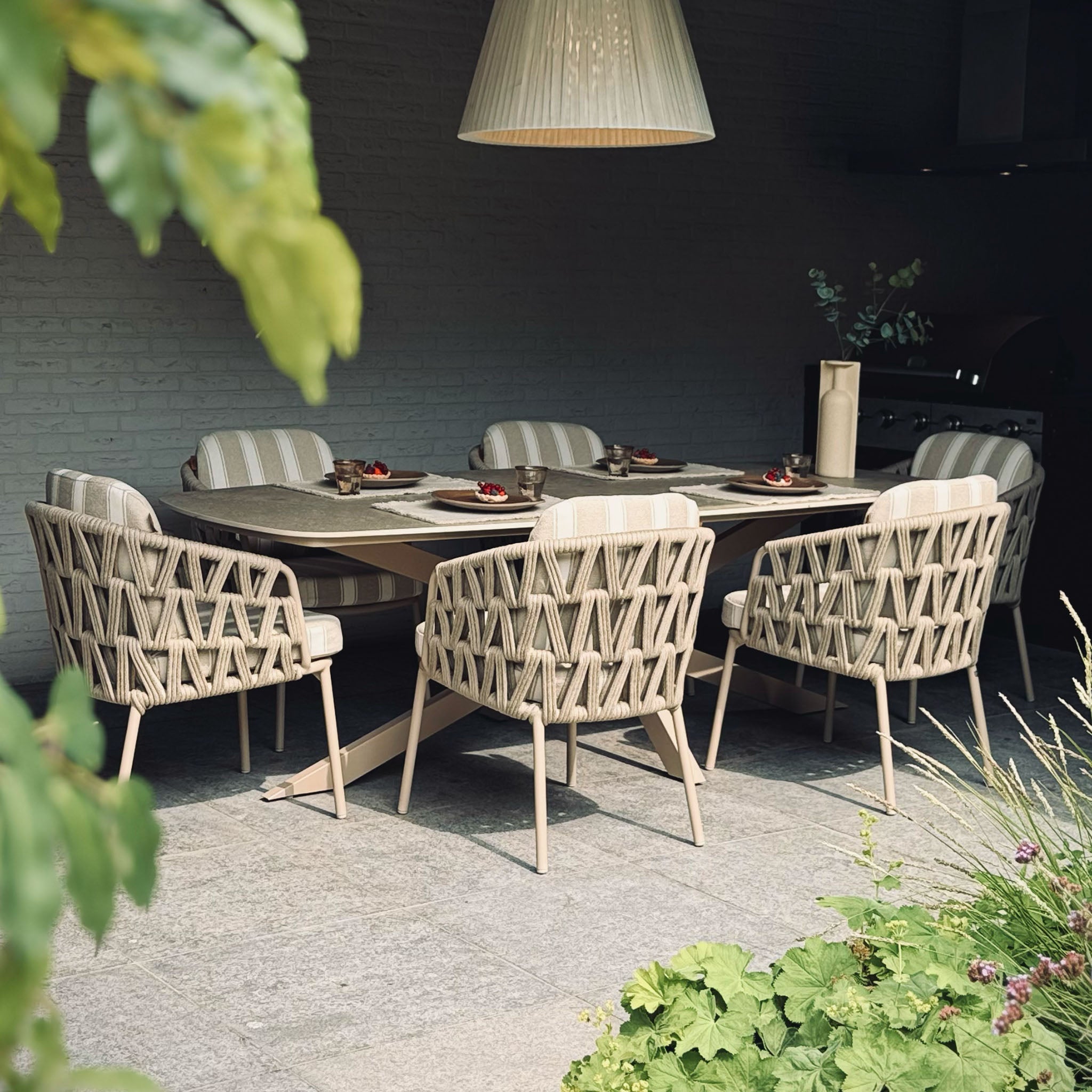 The Madeira 6 Seat Curved Rectangular Dining Set in Striped Latte features six curved chairs and a powder-coated aluminium table, perfect for outdoor dining under a large hanging light.