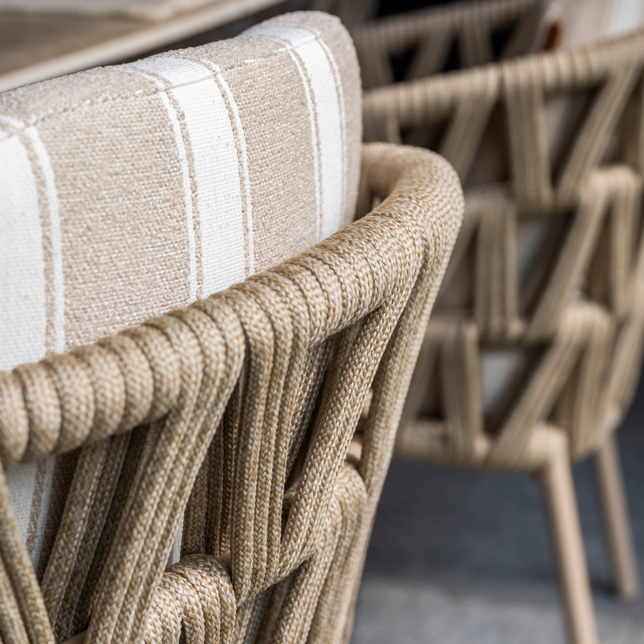 Close-up of the Madeira 6 Seat Curved Rectangular Dining Set in Striped Latte, featuring beige woven rope chairs with striped cushions and a powder-coated aluminium table on a gray floor for stylish outdoor dining.