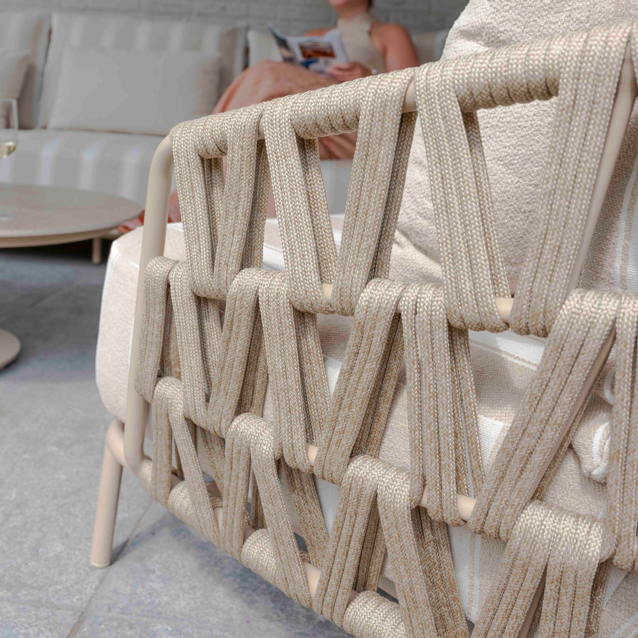 Close-up of a beige woven garden chair back with a blurred background of a woman reading—part of the elegant Madeira 3 Seat Sofa Set in Striped Latte.