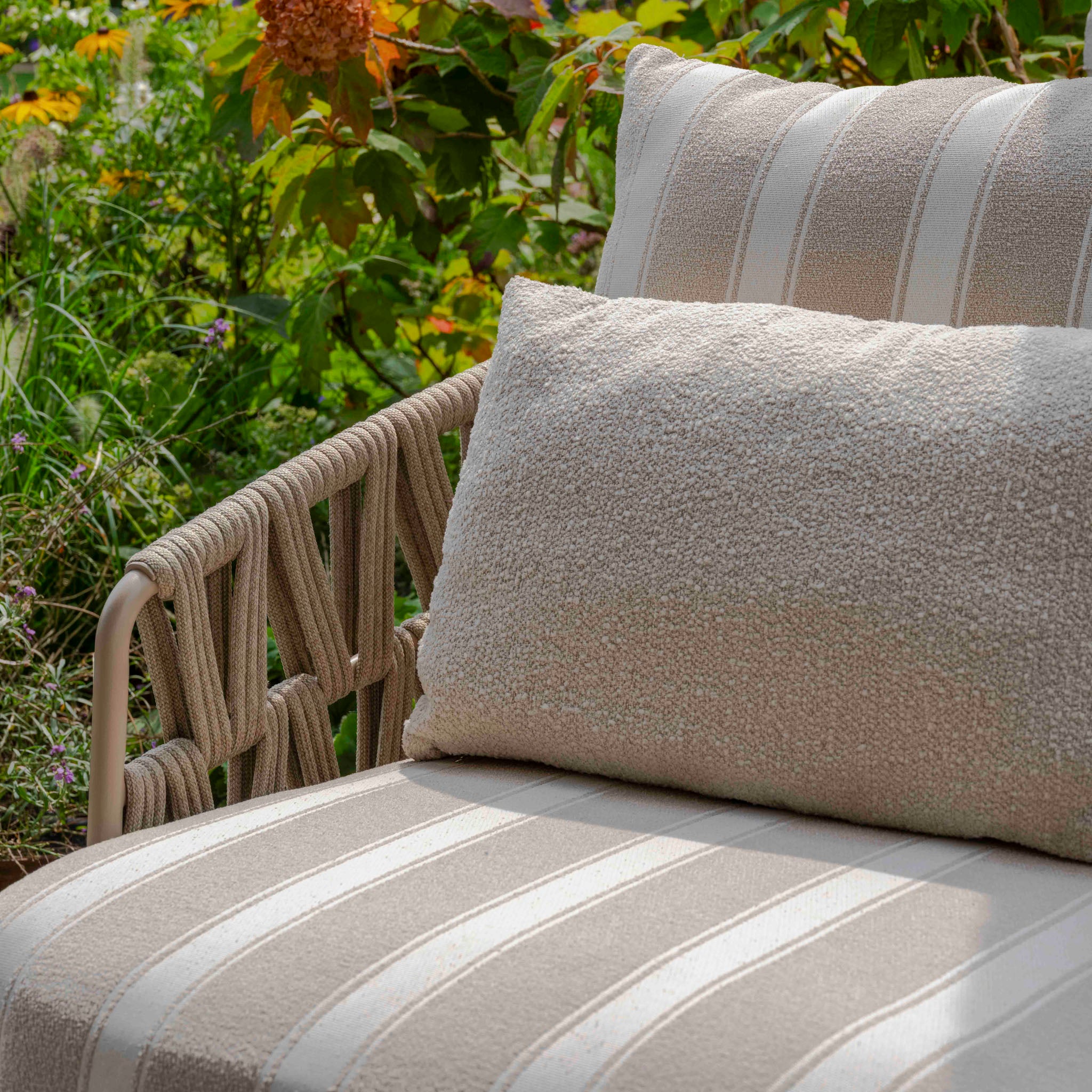 The Madeira 3 Seat Sofa Set in Striped Latte with cushions creates a cozy, stylish space among greenery and flowers—perfect for relaxing with elegant garden furniture.