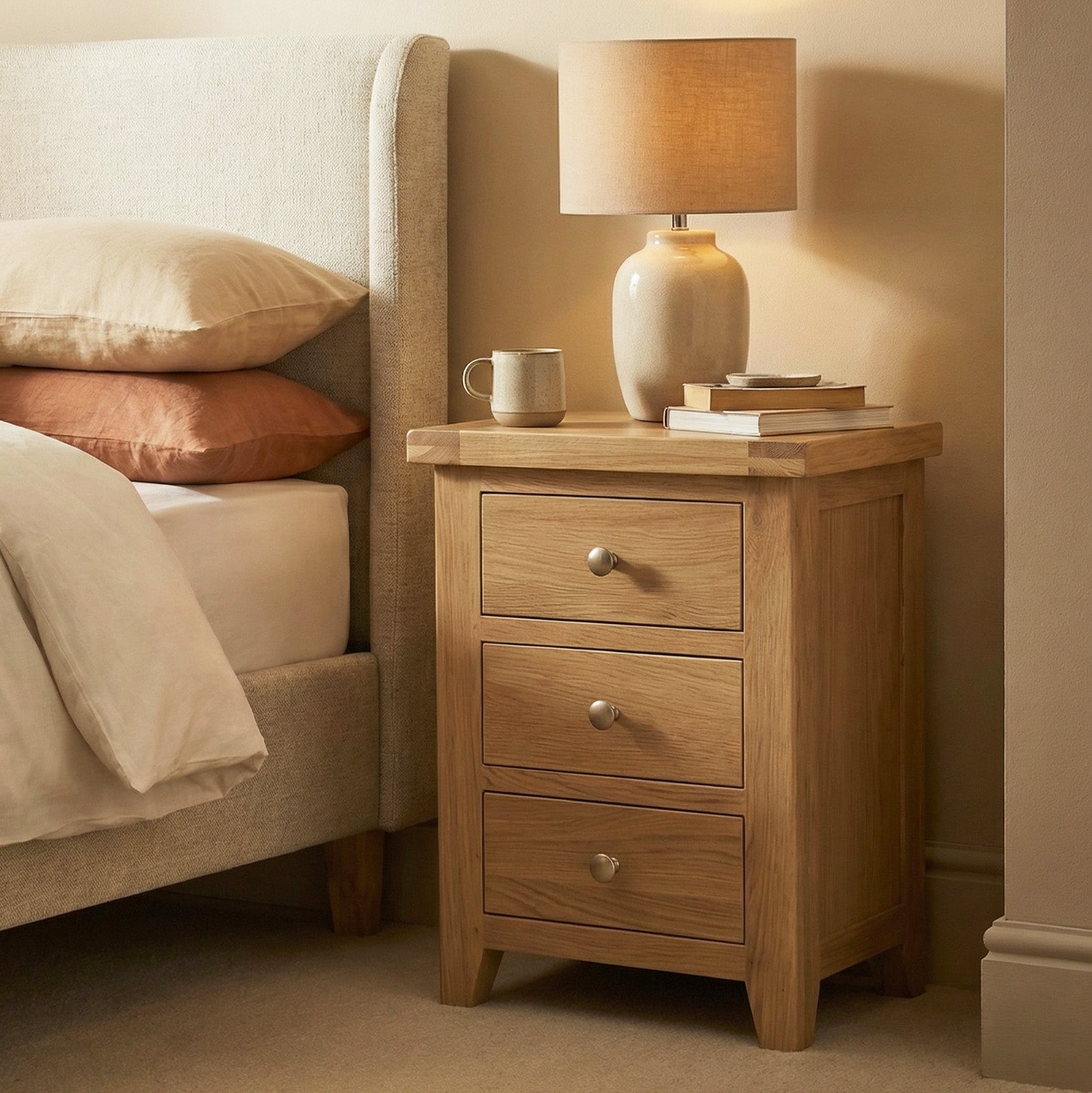 The Burford 3 Drawers Bedside Table in Natural Oak, topped with a lamp, books, and a mug, sits beside a neatly made bed.