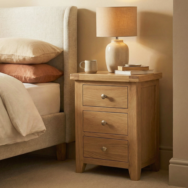The Burford 3 Drawers Bedside Table in Natural Oak, topped with a lamp, books, and a mug, sits beside a neatly made bed.