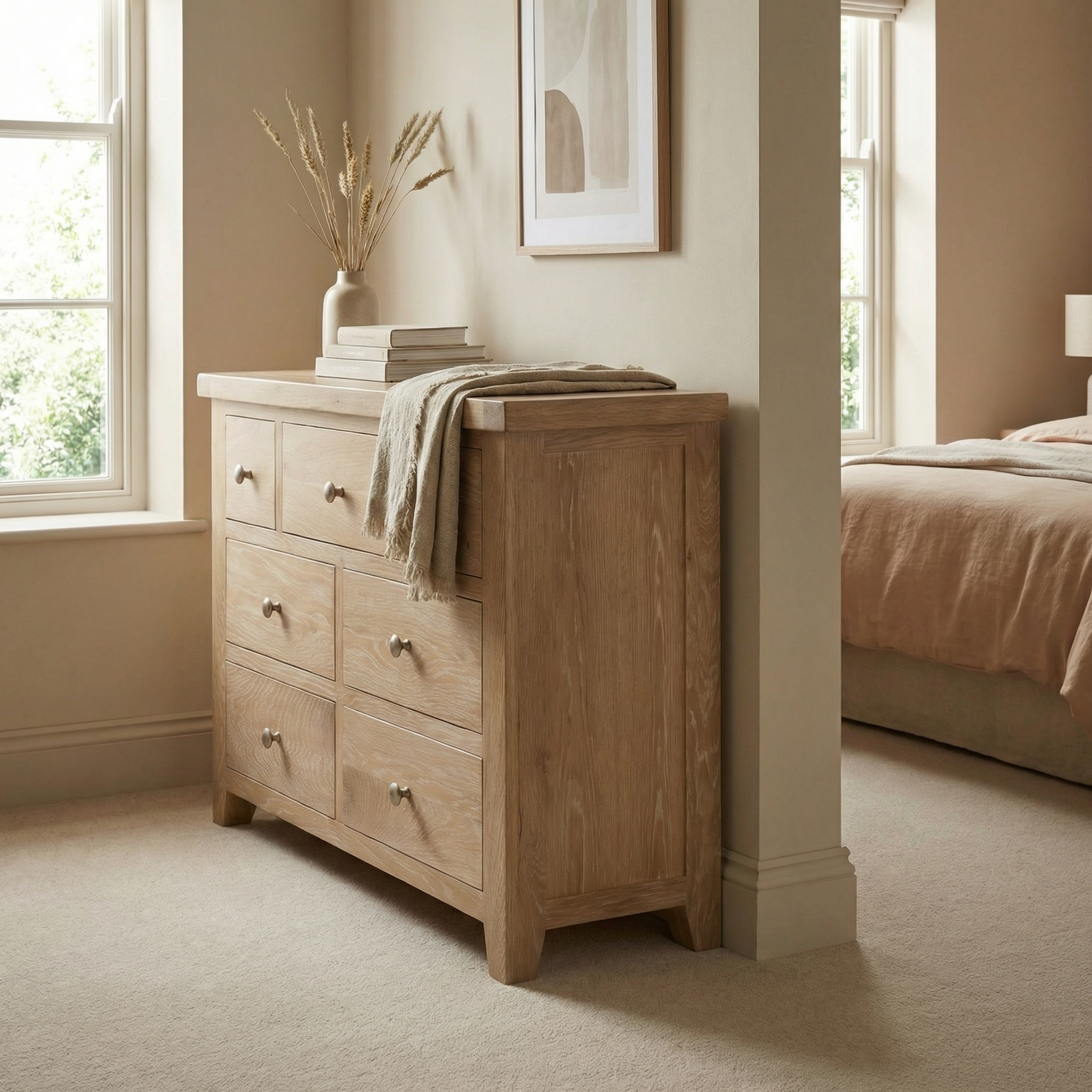 The Burford 3 Over 4 Chest of Drawers in Natural Oak stands as a timeless storage piece, complementing a beige bedroom with books and a vase while basking in soft natural light.