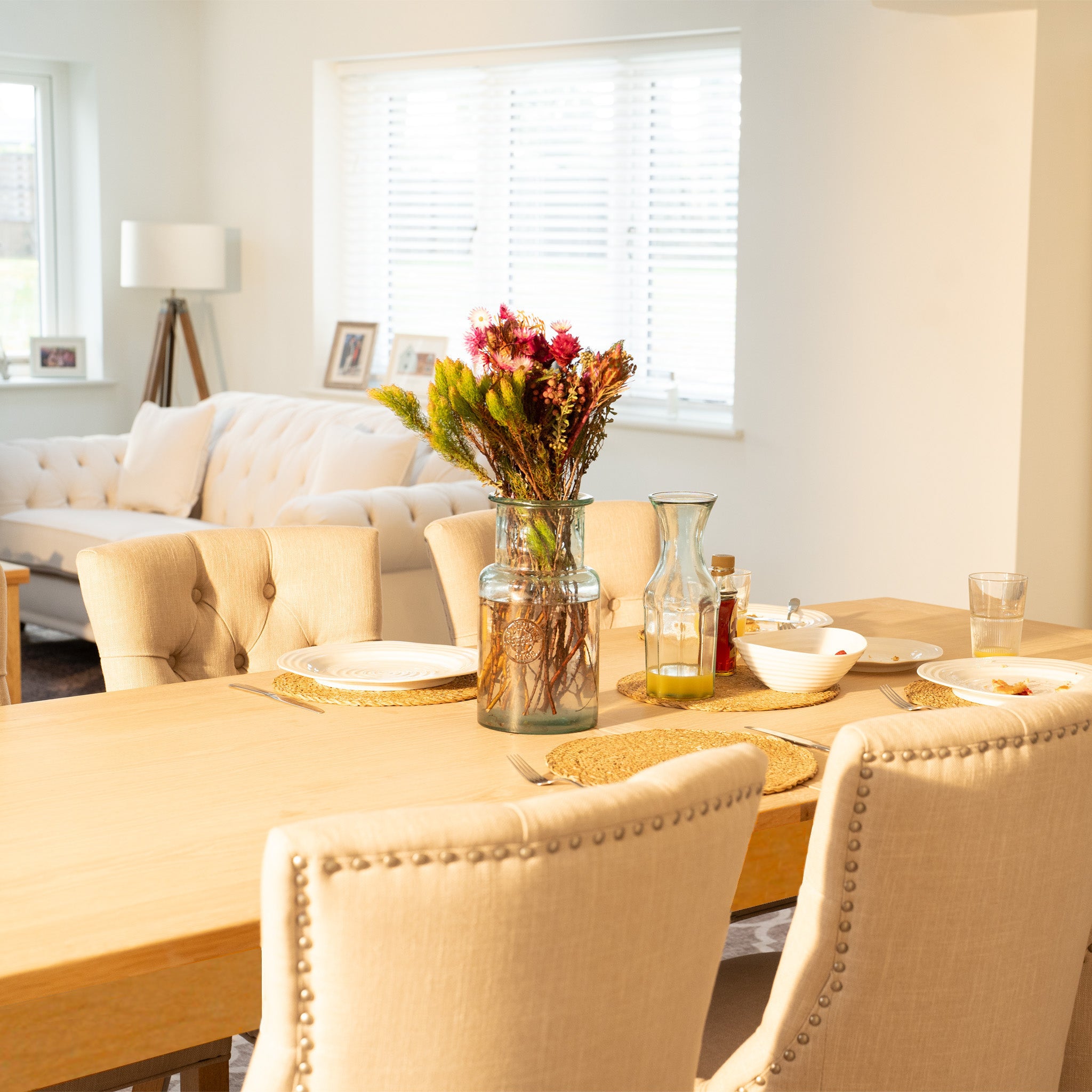 The Burford 8 Seat Extending Dining Set with Regent Chairs in Natural Oak and Ivory, complete with tableware and a vase of flowers, enhances this bright, modern living space.