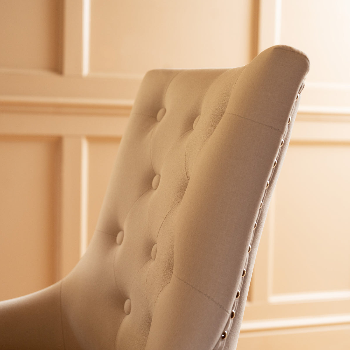 Close-up of a beige tufted Regent chair with button details against a light-colored paneled wall, perfectly complementing the timeless elegance of the Burford 8 Seat Extending Dining Set in Natural Oak and Ivory.