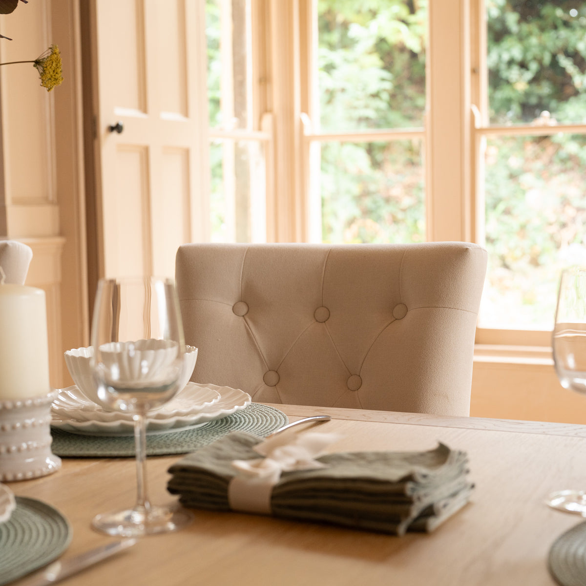 The Burford 8 Seat Extending Dining Set with Regent Chairs in Natural Oak and Ivory is beautifully arranged with plates, glasses, folded napkins, and a cushioned chair by sunny windows.